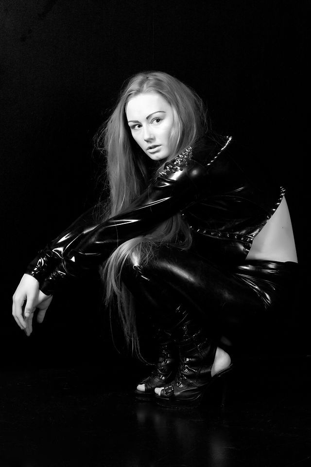 Black and white photo of a woman in shiny latex outfit crouching against dark background.