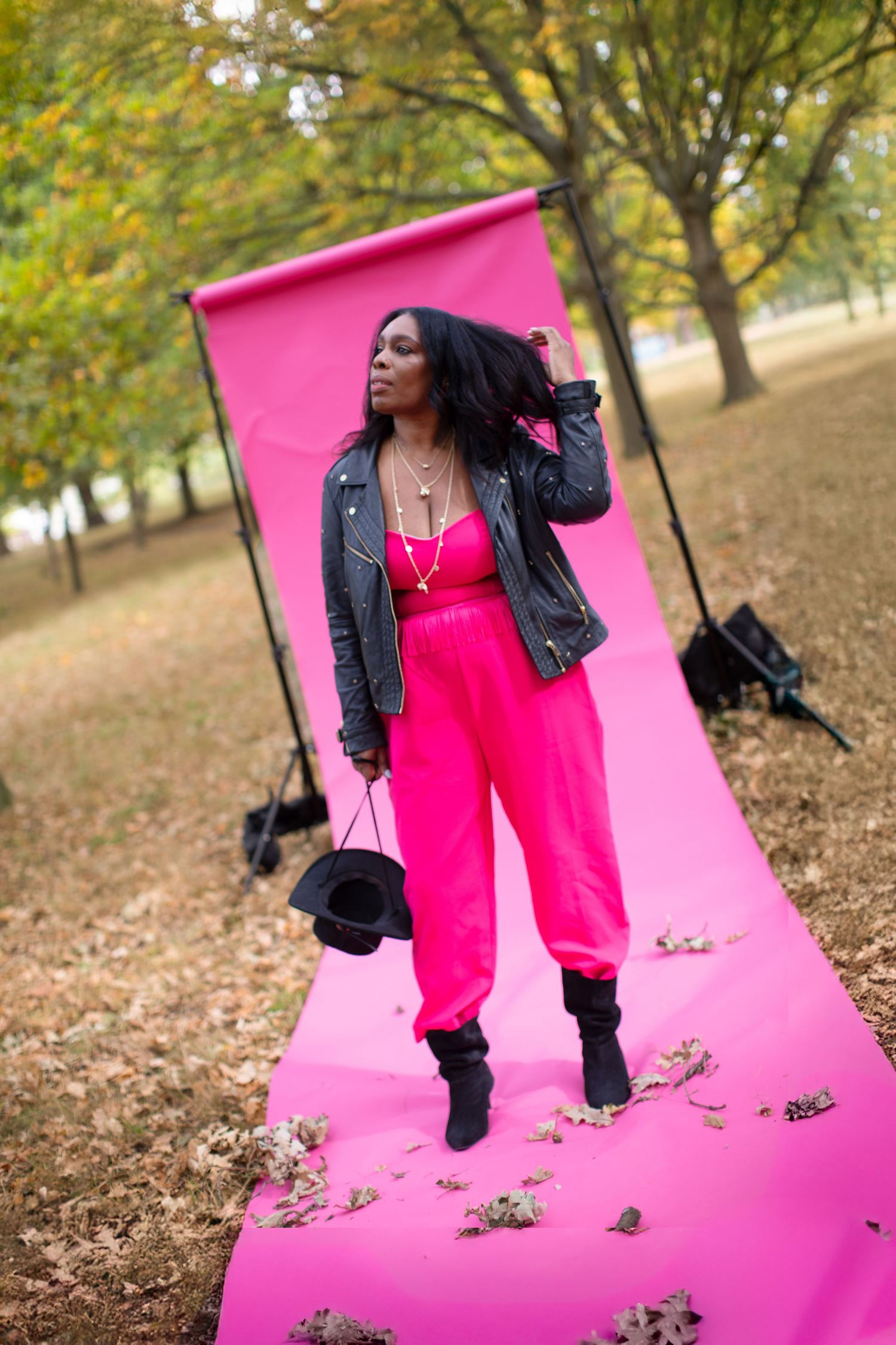 A woman in a pink jumpsuit and leather jacket poses on a pink backdrop in an autumn park.