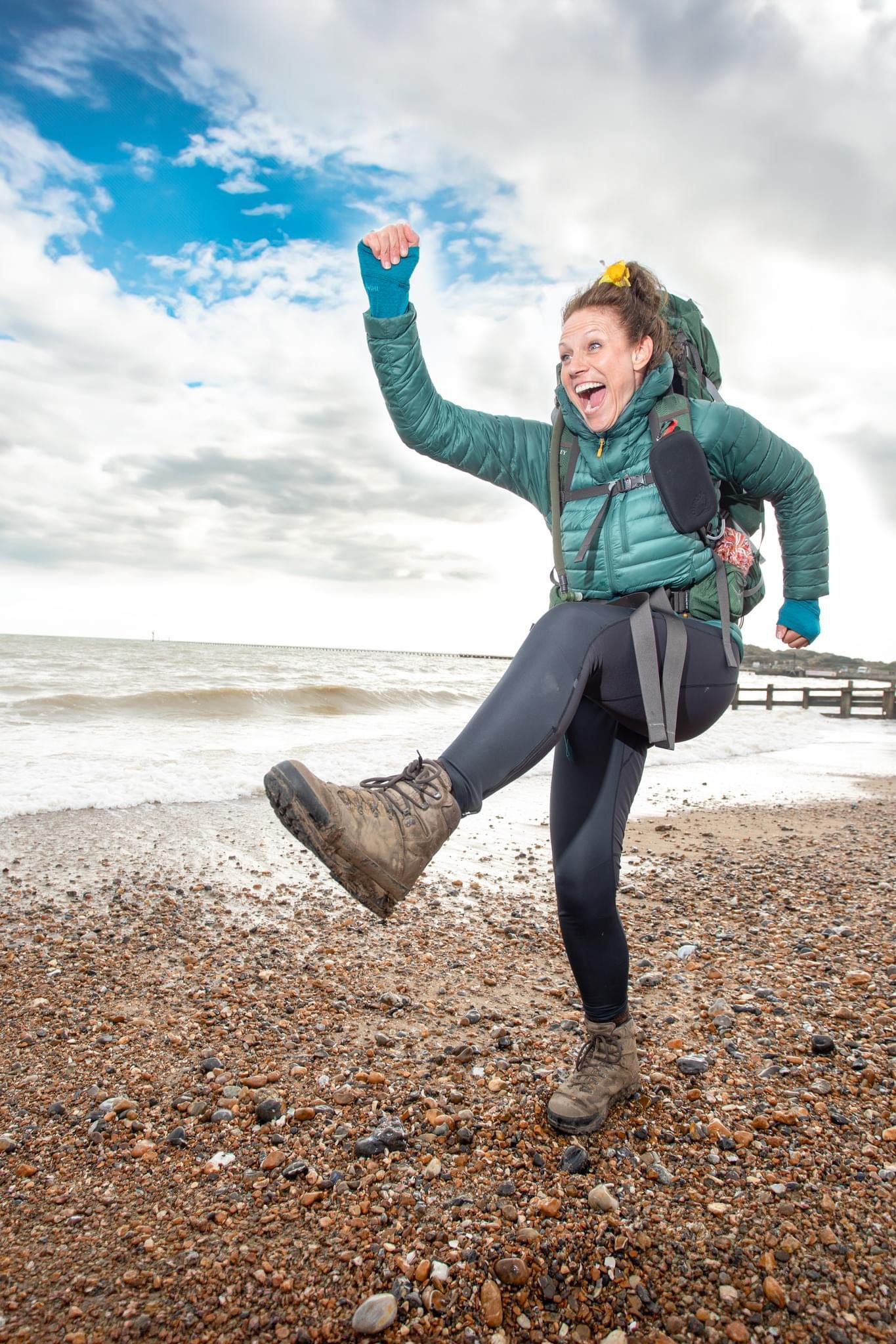 A cheerful woman hiker with a backpack celebrates on a pebble beach, raising her fist and kicking her leg.