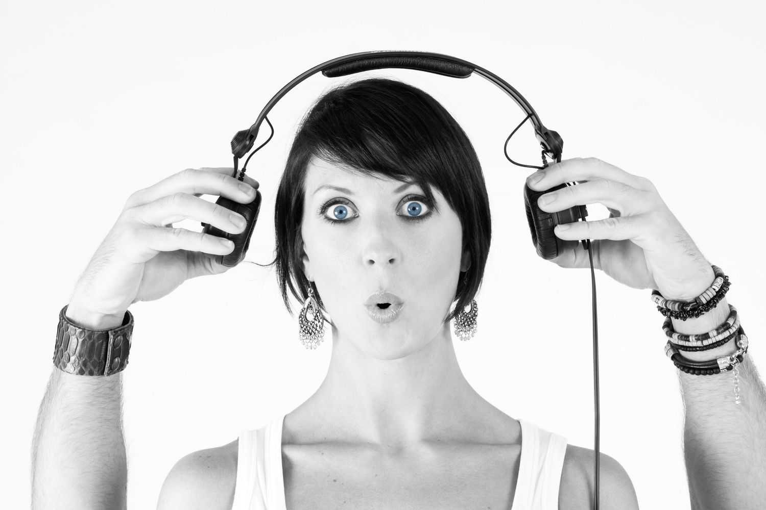 Black and white photo of a surprised woman with short dark hair holding large headphones, wearing bracelets and a tank top.
