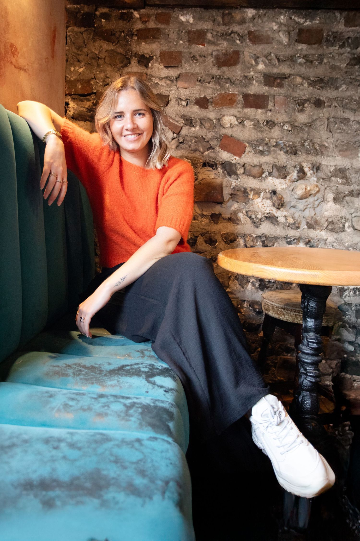 Person relaxing on a teal velvet couch wearing an orange sweater in a rustic brick-walled setting.