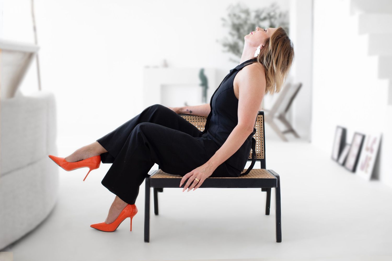 Woman in black outfit sits on bench with bright orange heels in minimalist white setting.