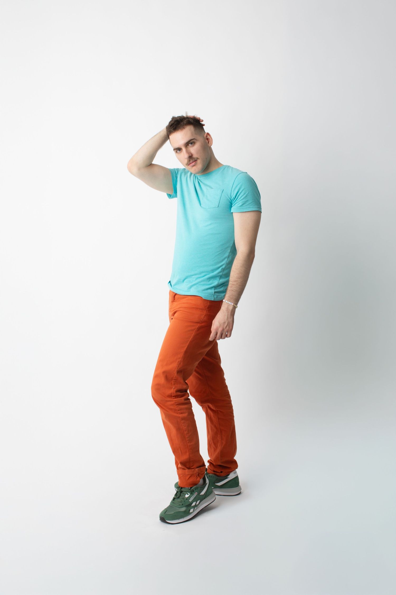 Man posing in teal t-shirt, rust orange pants, and green sneakers against white background.