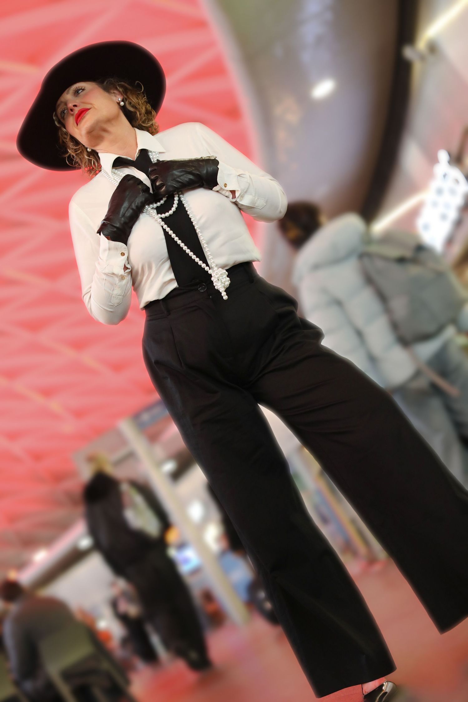 Elegant woman in black wide-leg pants, white shirt, pearl necklace, black hat and gloves poses at a fashion event.