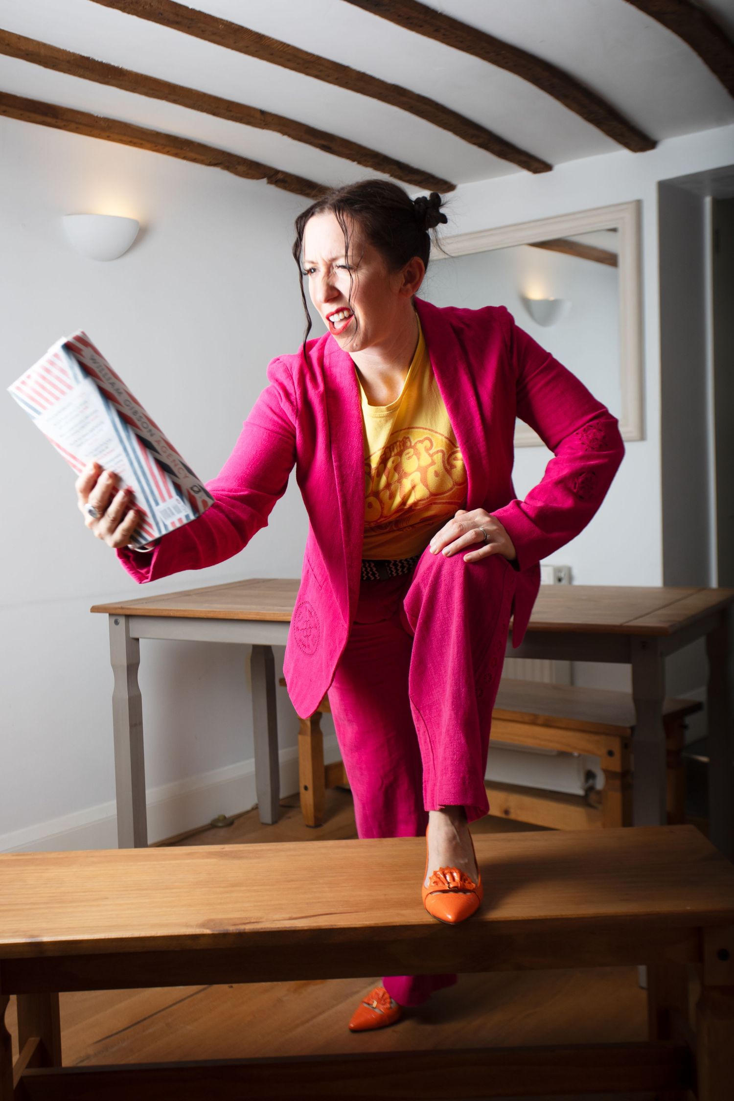 Woman in pink suit holding a magazine while standing on a wooden bench in a rustic interior.