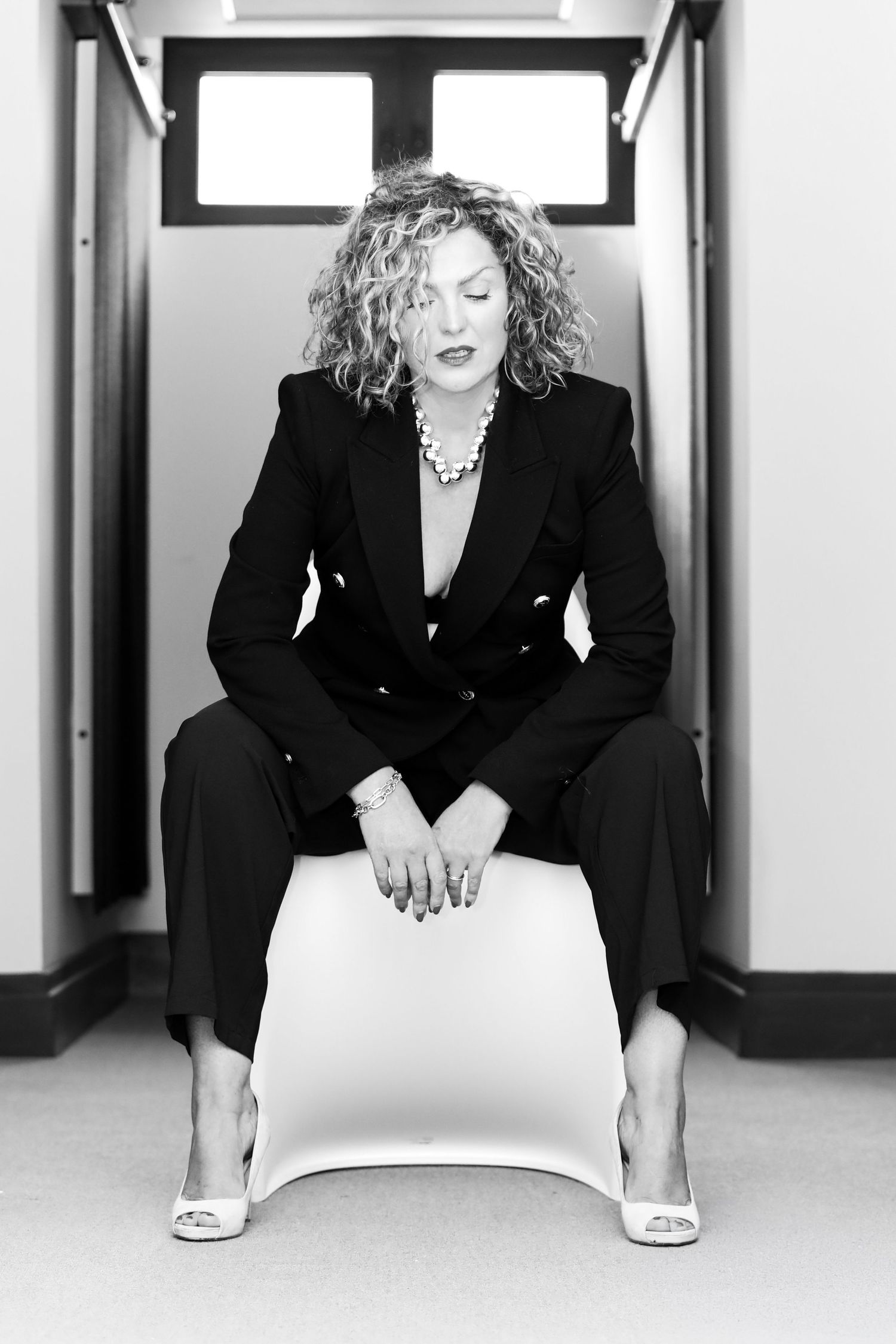 Black and white photo of a confident woman in a black suit sitting on a white stool in a hallway.