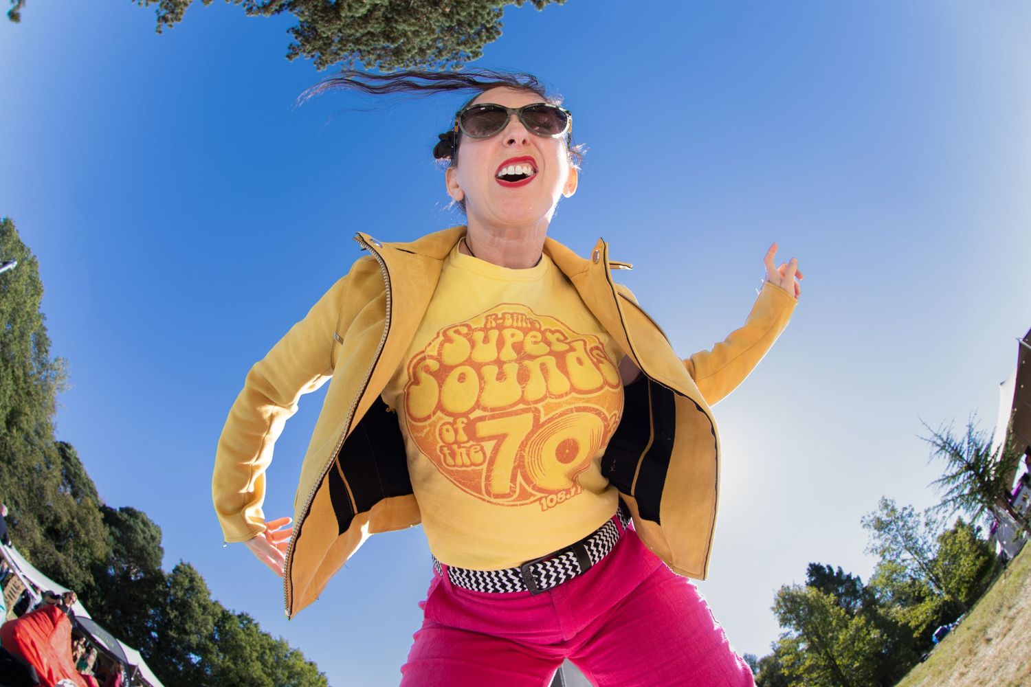 Woman in yellow jacket and pink pants dancing outdoors at a festival, wearing sunglasses and a retro 70s graphic tee.