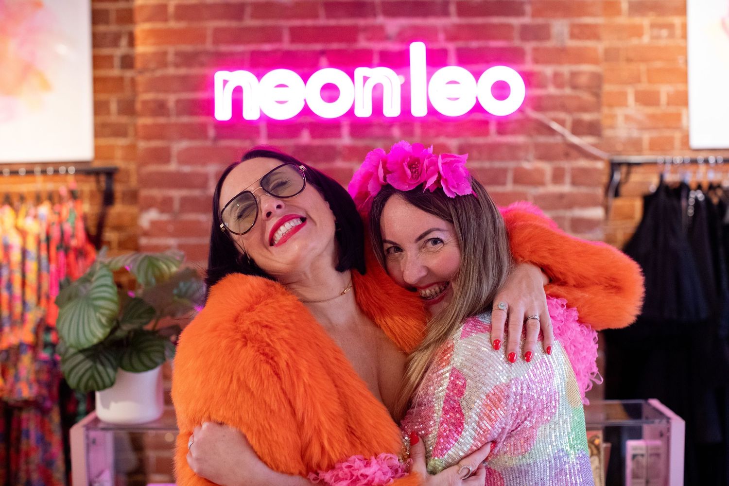 Two women hugging and smiling in front of a neon Neon Leo sign in a colorful boutique clothing store.