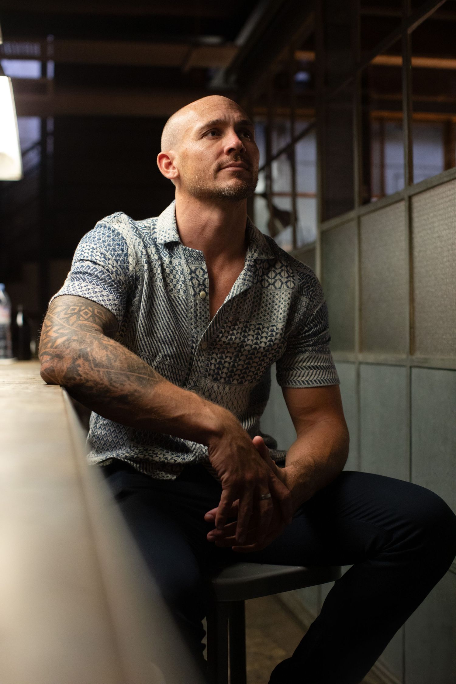 Bald tattooed muscular man in patterned polo shirt sitting thoughtfully in a dimly lit industrial space.