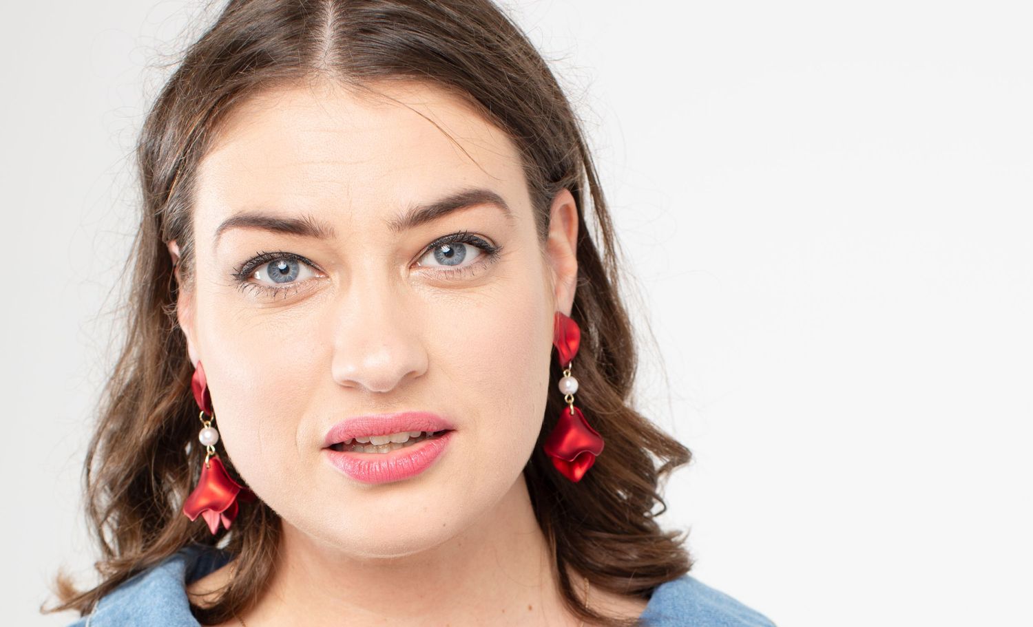 A young woman with blue eyes wearing bold red drop earrings poses against a white background.