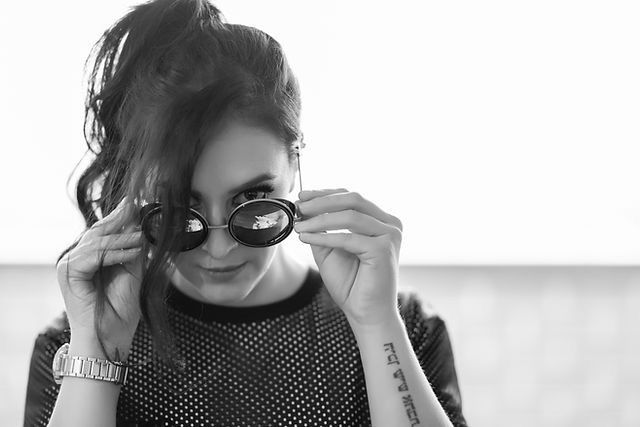 Black and white photo of a young woman with sunglasses, tattoo on arm, wearing a mesh top, looking at camera.
