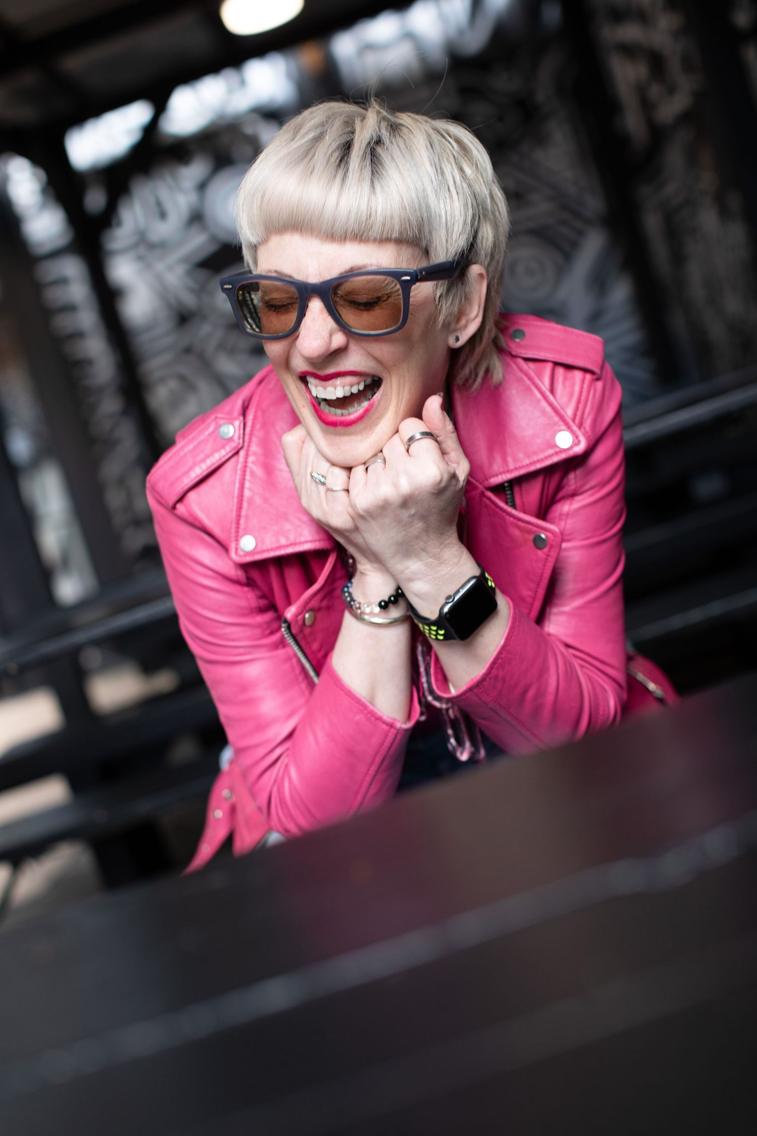 Woman in pink leather jacket laughing joyfully while wearing stylish sunglasses outdoors.