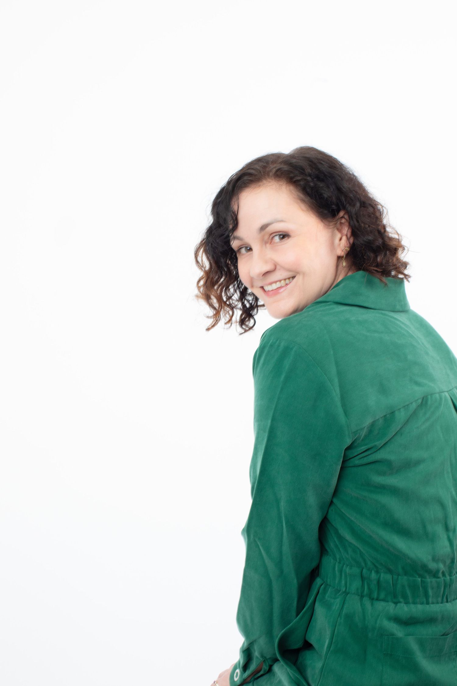 Smiling woman with curly dark hair wearing a green jacket, looking over her shoulder against a white background.
