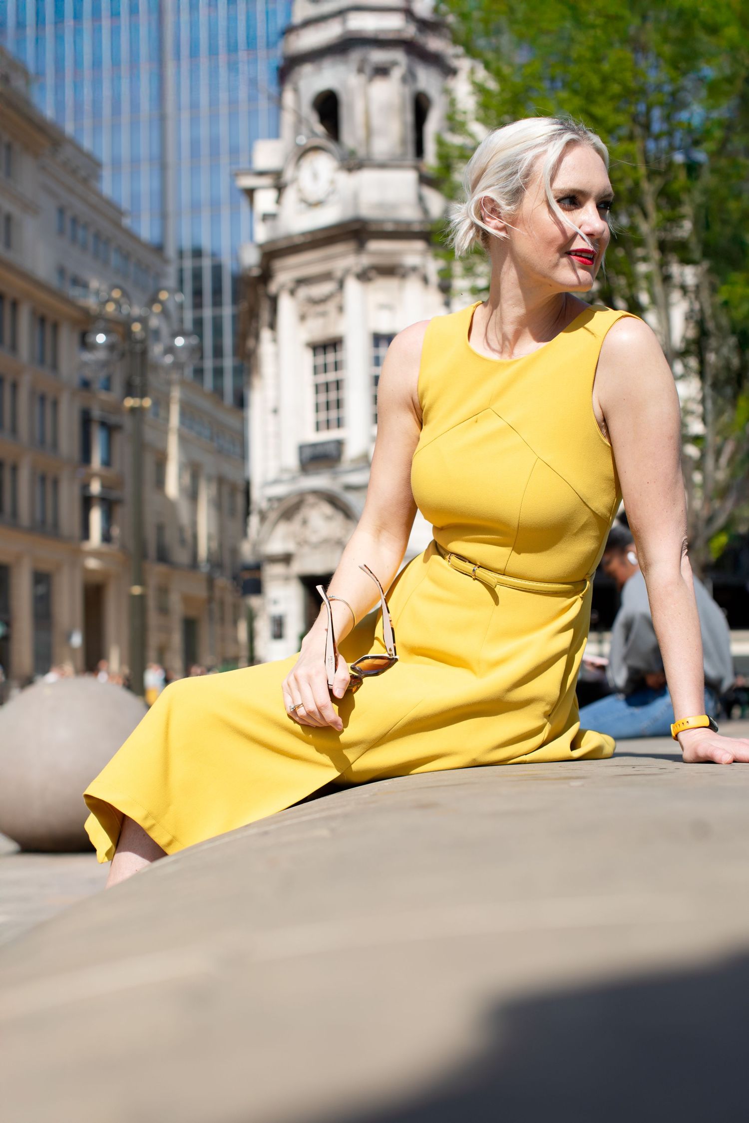 Person in yellow dress sitting on stone ledge with classic architecture in background.