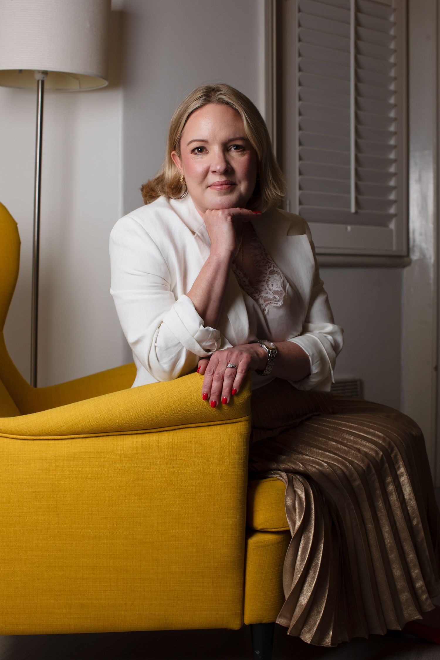 Blonde woman in white blazer and gold skirt sits relaxed in yellow armchair, resting chin on hand.