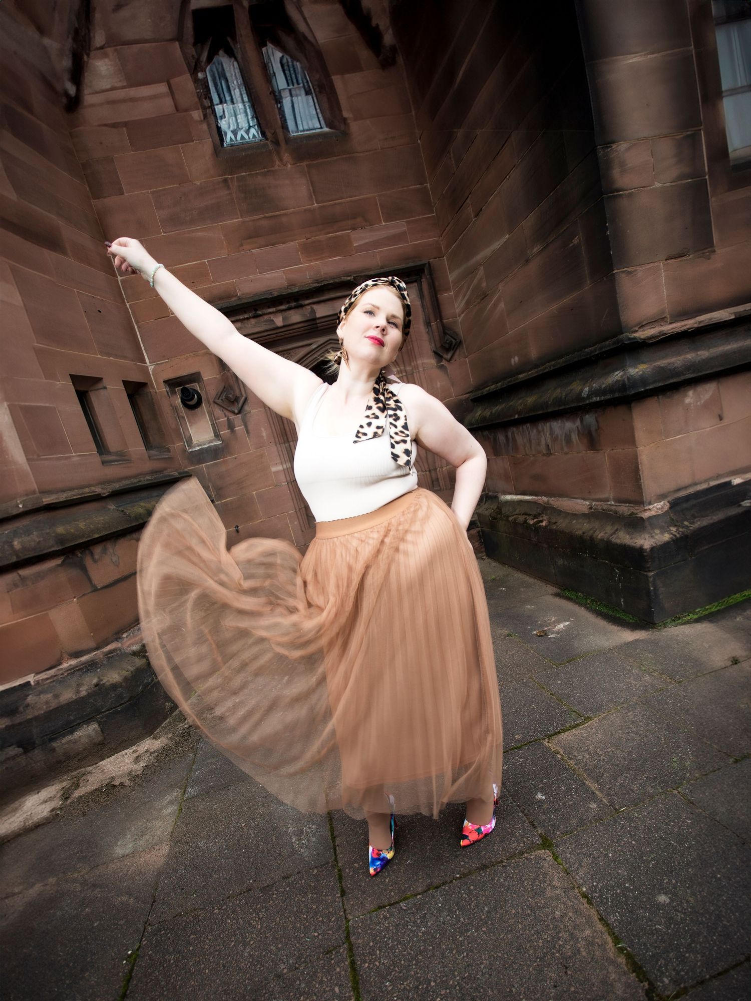 Woman in white crop top, leopard scarf, tan tulle skirt, and colorful heels posing gracefully outdoors near a stone building.