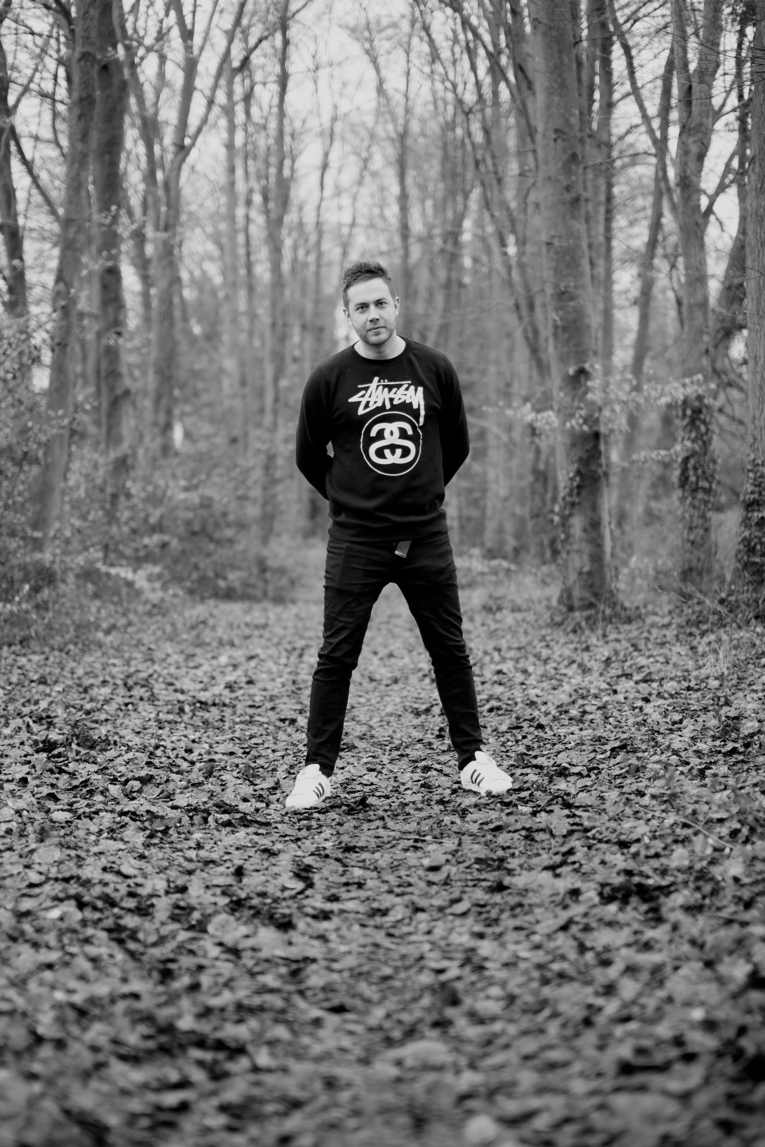 Black and white forest portrait of person in Stussy sweatshirt standing among bare trees.