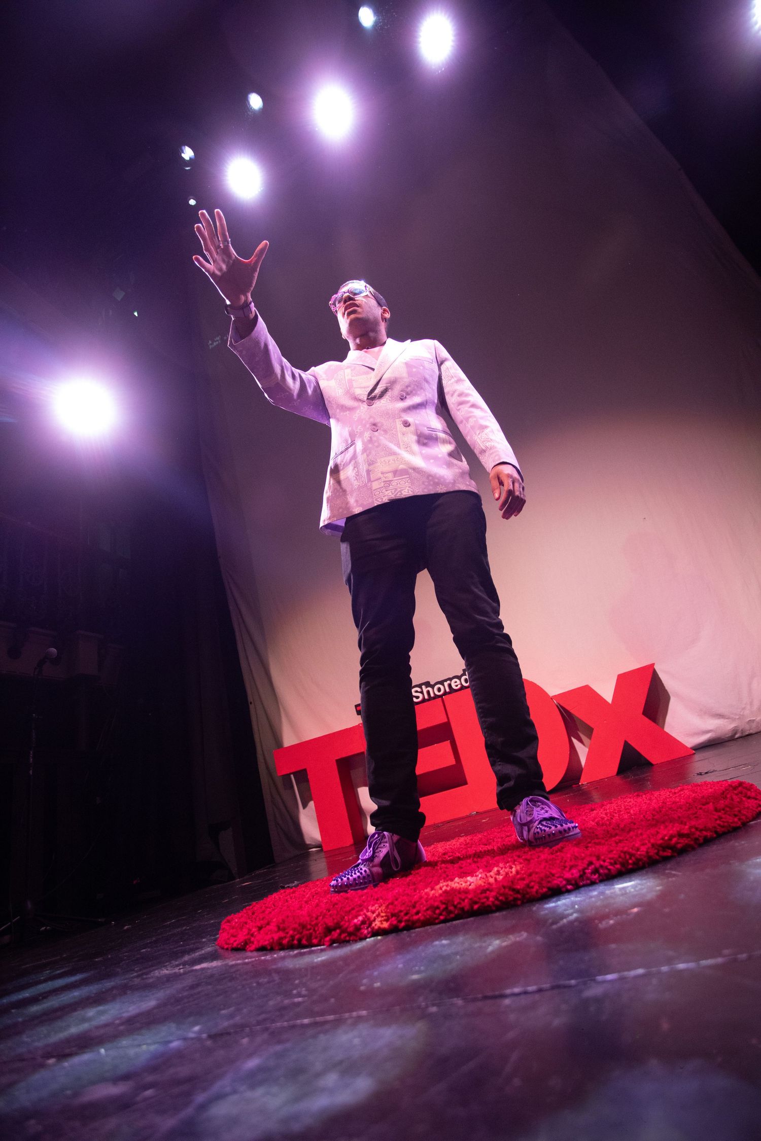 Man in white blazer gesturing on a TEDx stage under dramatic purple lighting.
