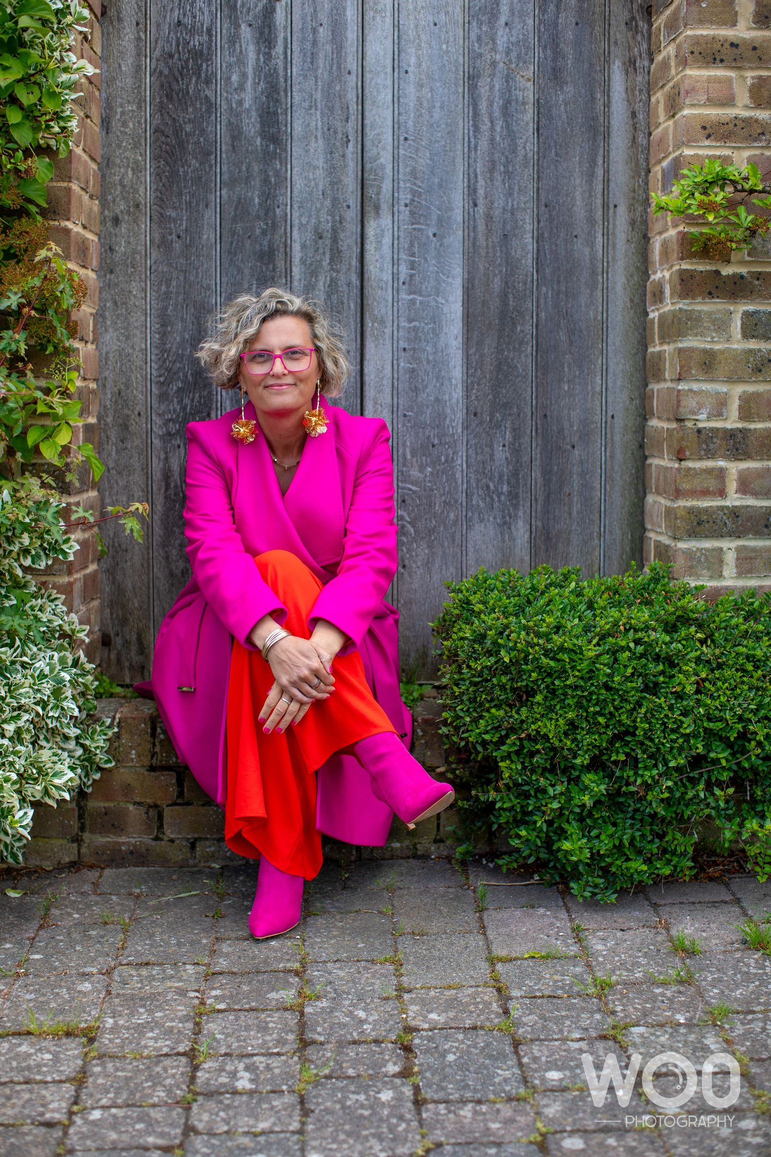 Fashion shoot featuring vibrant hot pink blazer and red dress against weathered wooden fence.
