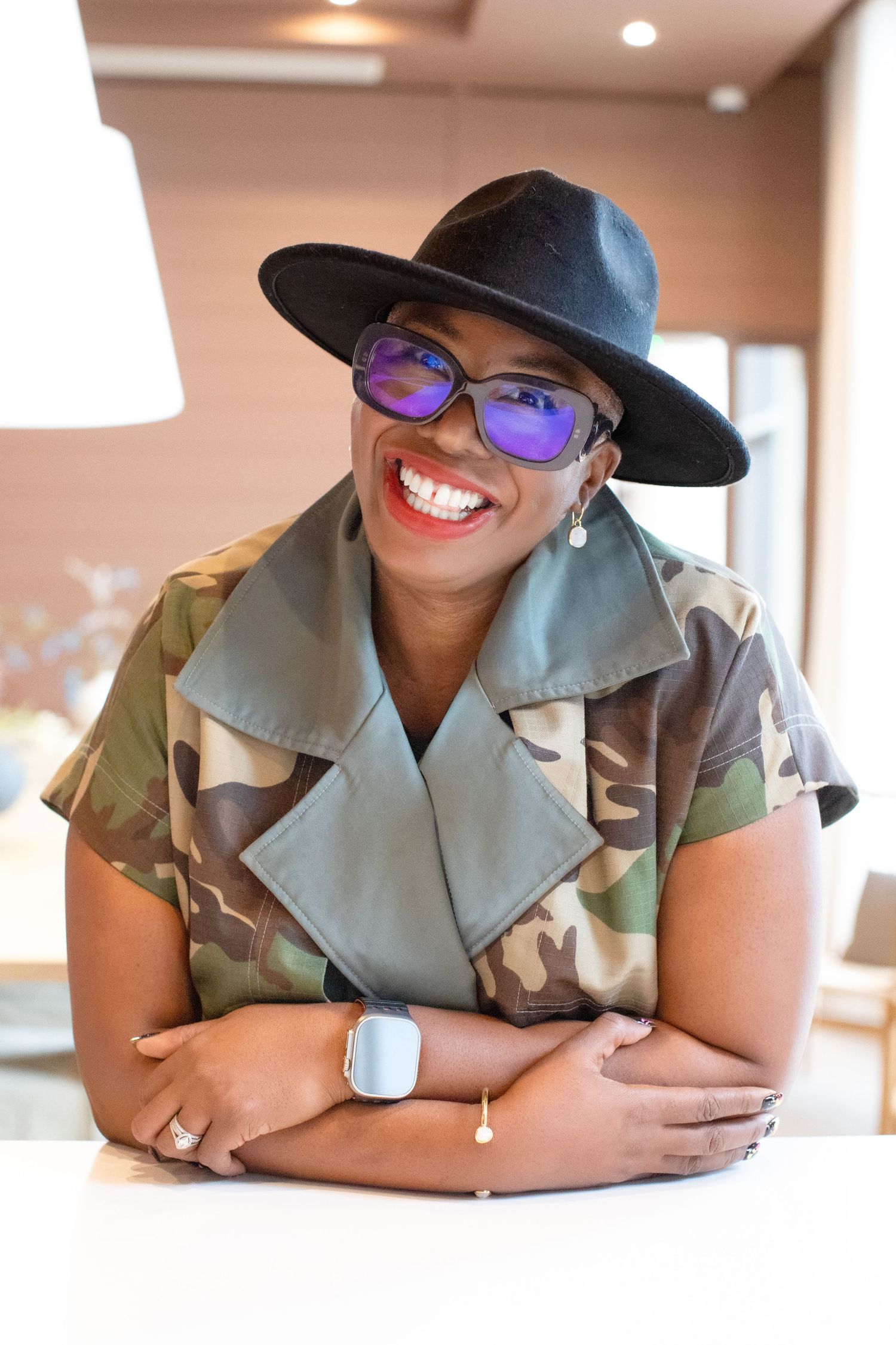 A stylish woman wearing purple mirrored sunglasses and a black hat smiles while sitting at a white table.