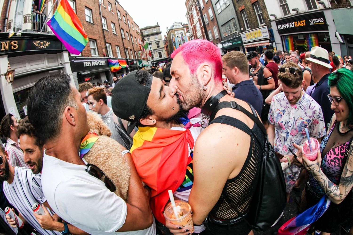 Two people kissing at a vibrant Pride parade surrounded by colorful crowds and rainbow flags on a busy street.