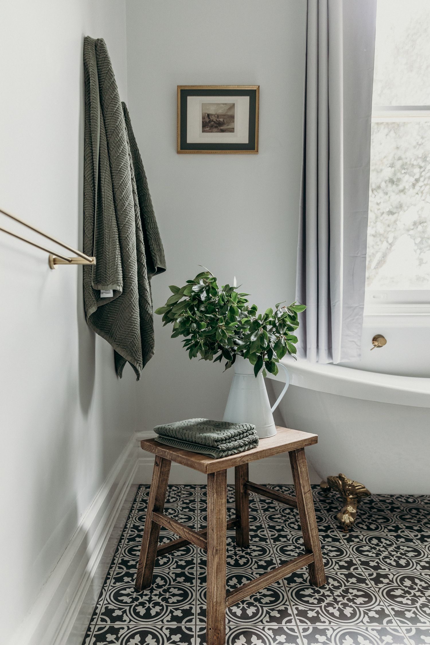 Minimalist bathroom corner with wooden stool, green towels, and potted plant on patterned tile floor.