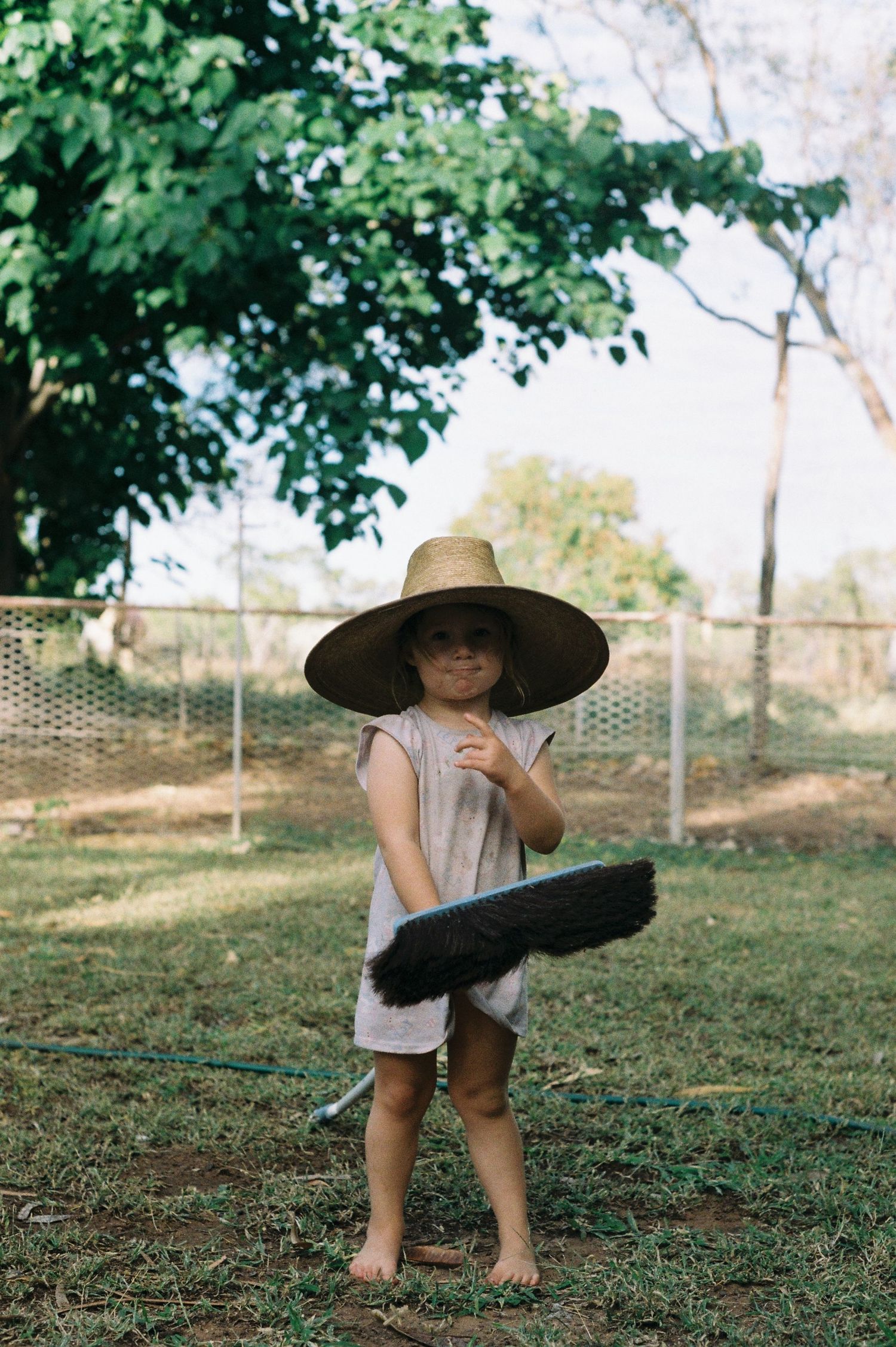 Child in wide-brimmed hat holding a broom in backyard setting.