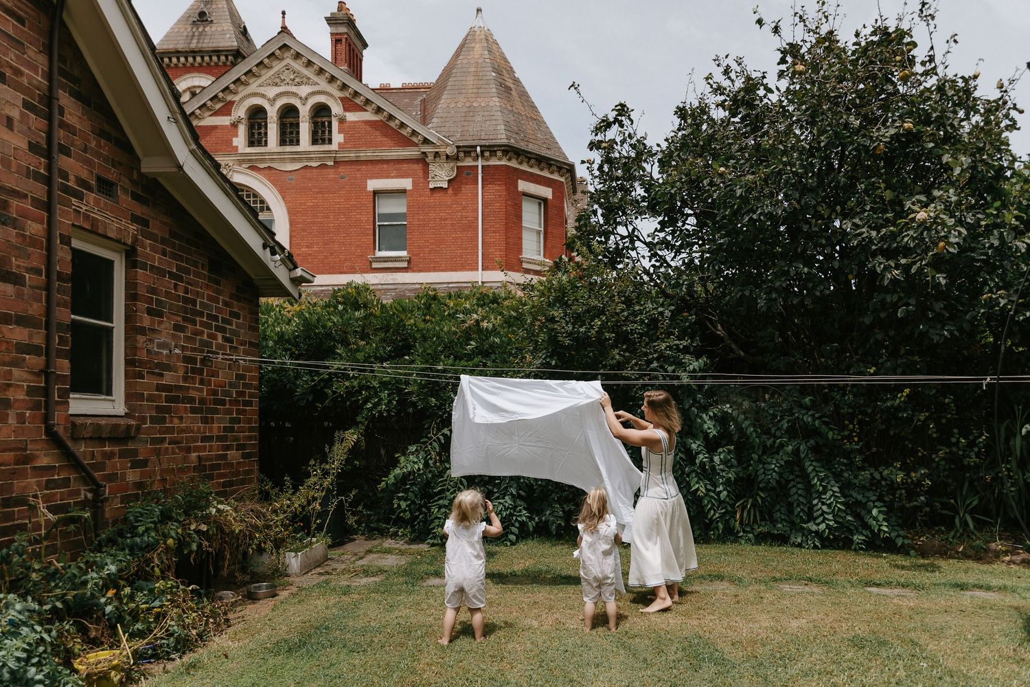 Children playing under a white sheet hung between trees in a garden with a historic brick house backdrop.