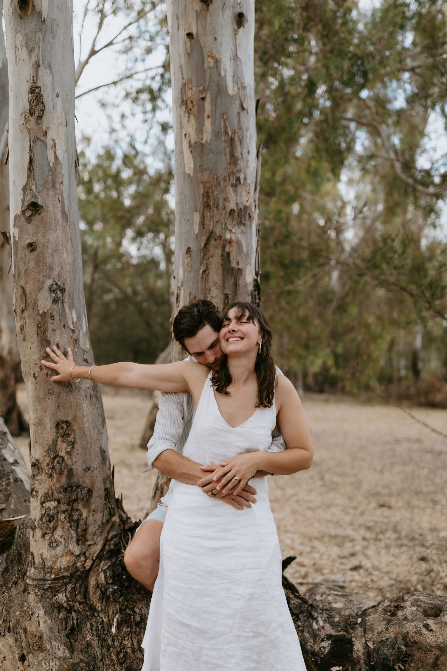 A couple shares intimate moments together while sitting among tall eucalyptus trees in a natural outdoor setting.