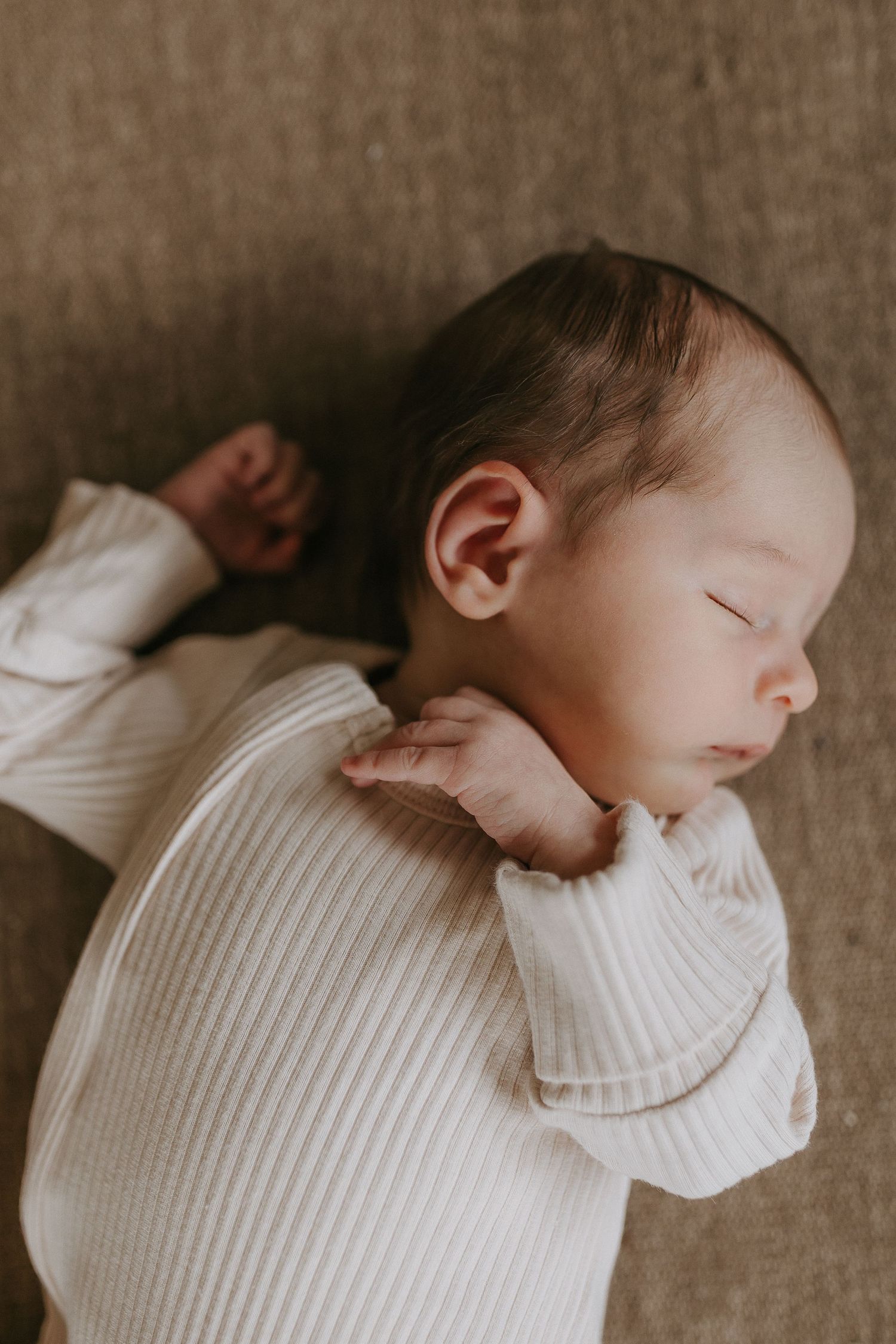 Sleeping newborn in white ribbed outfit rests peacefully on textured fabric.