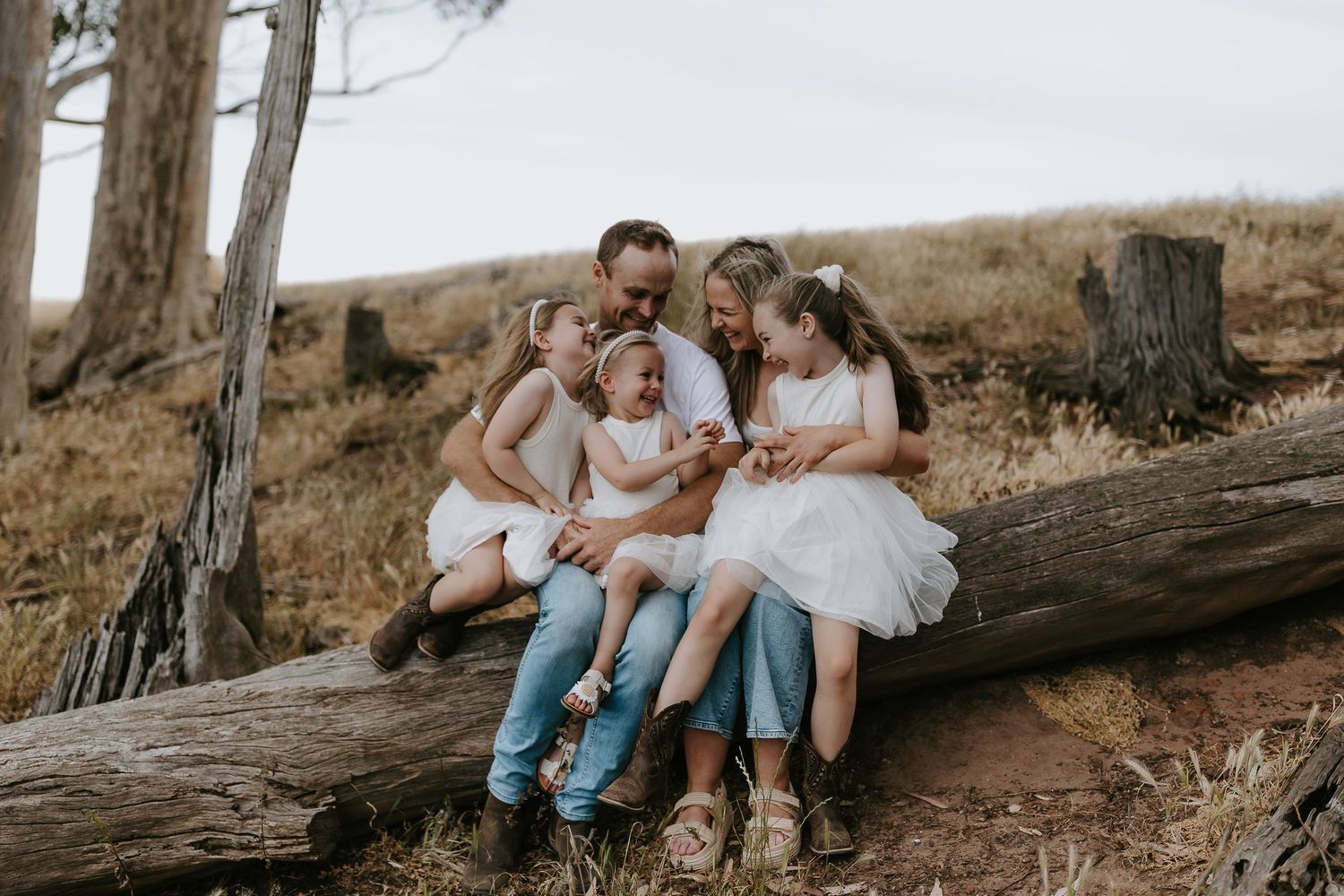 Family group relaxing on fallen tree trunk in wooded outdoor setting.