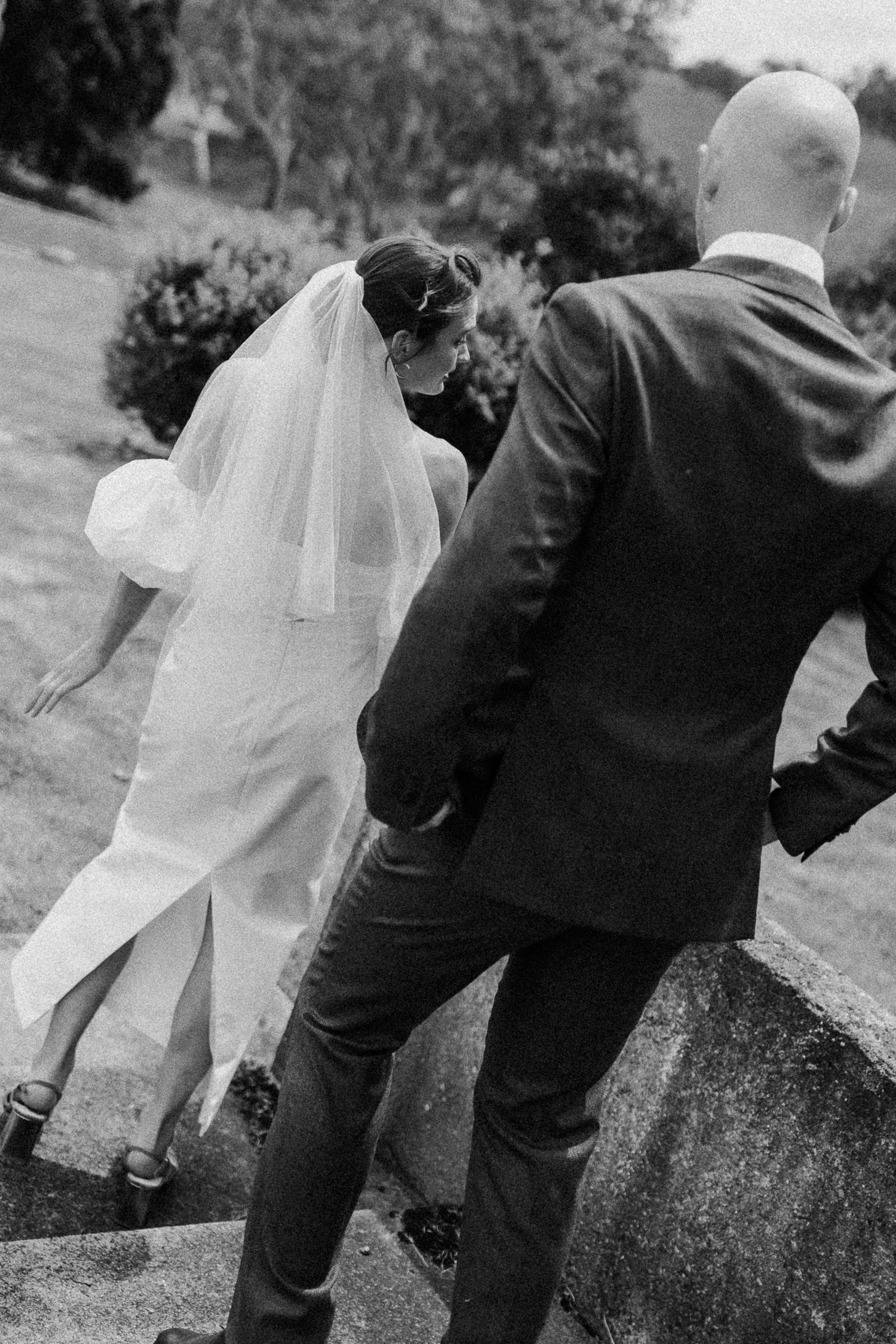 Black and white photo of a bride in flowing veil and groom walking together outdoors on their wedding day.