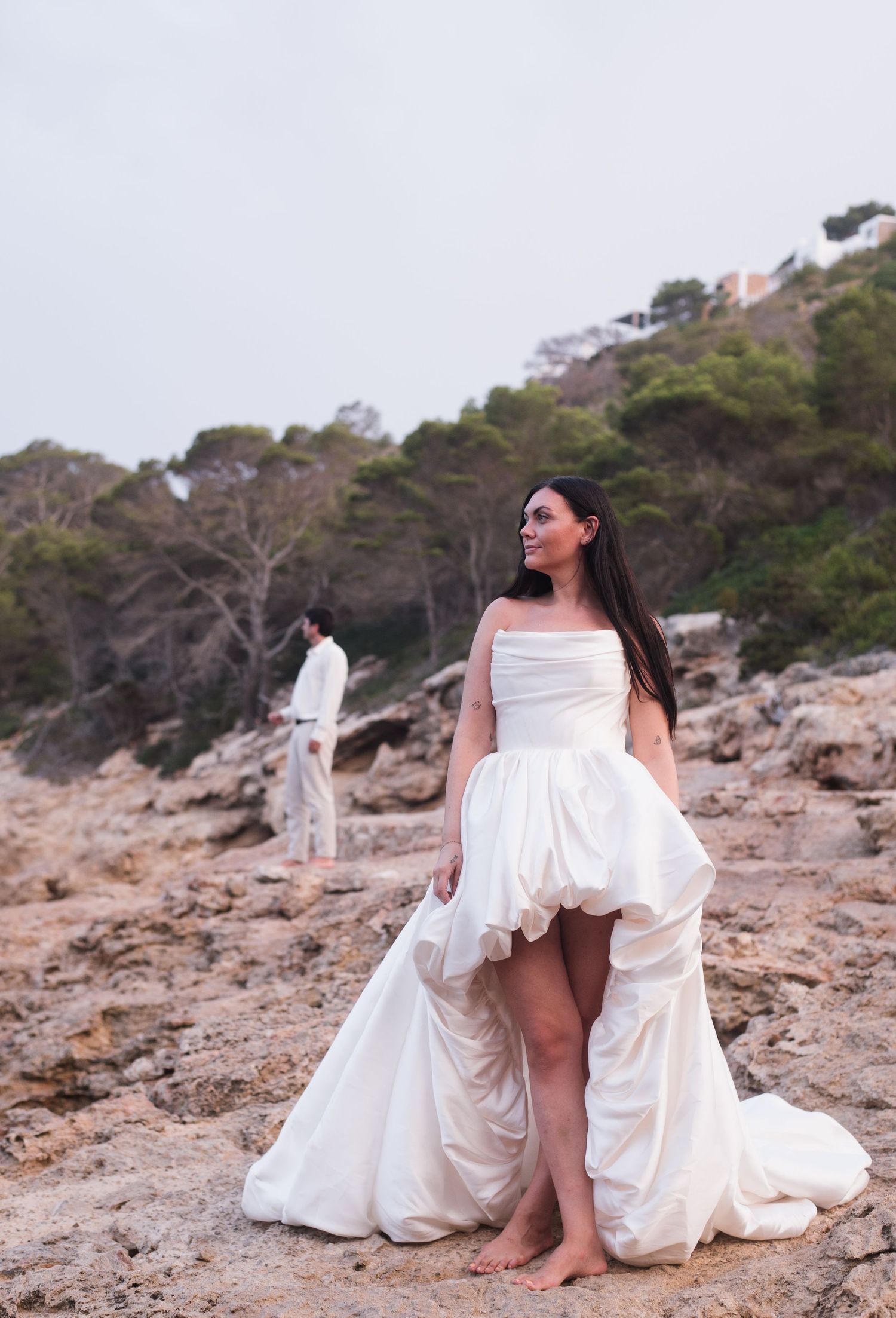 Bride in a flowing white strapless gown stands barefoot on rocky coastal terrain with groom in background.