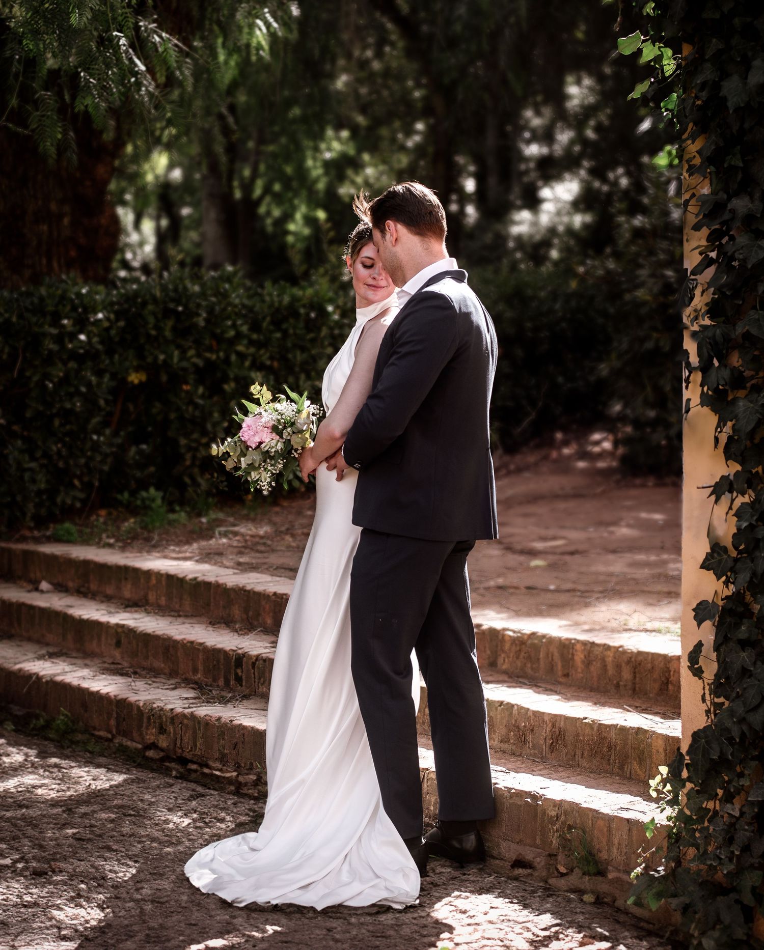 Bride and groom embracing on stone steps surrounded by lush greenery, bride holding a pink and white bouquet.