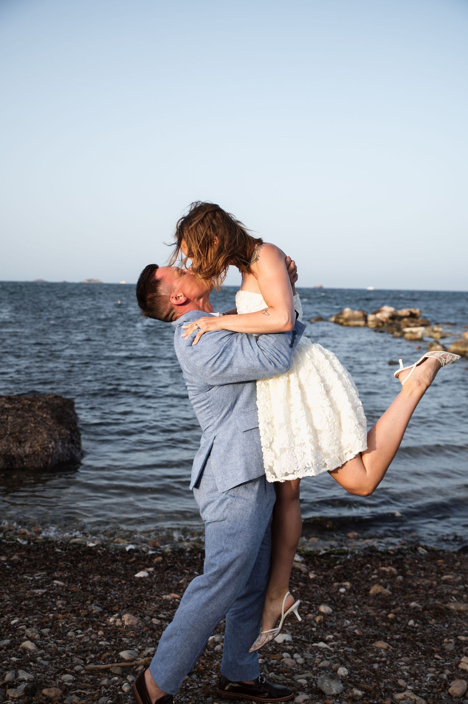 Couple sharing a romantic kiss on a rocky beach shore, man lifting woman in white lace dress, ocean backdrop.