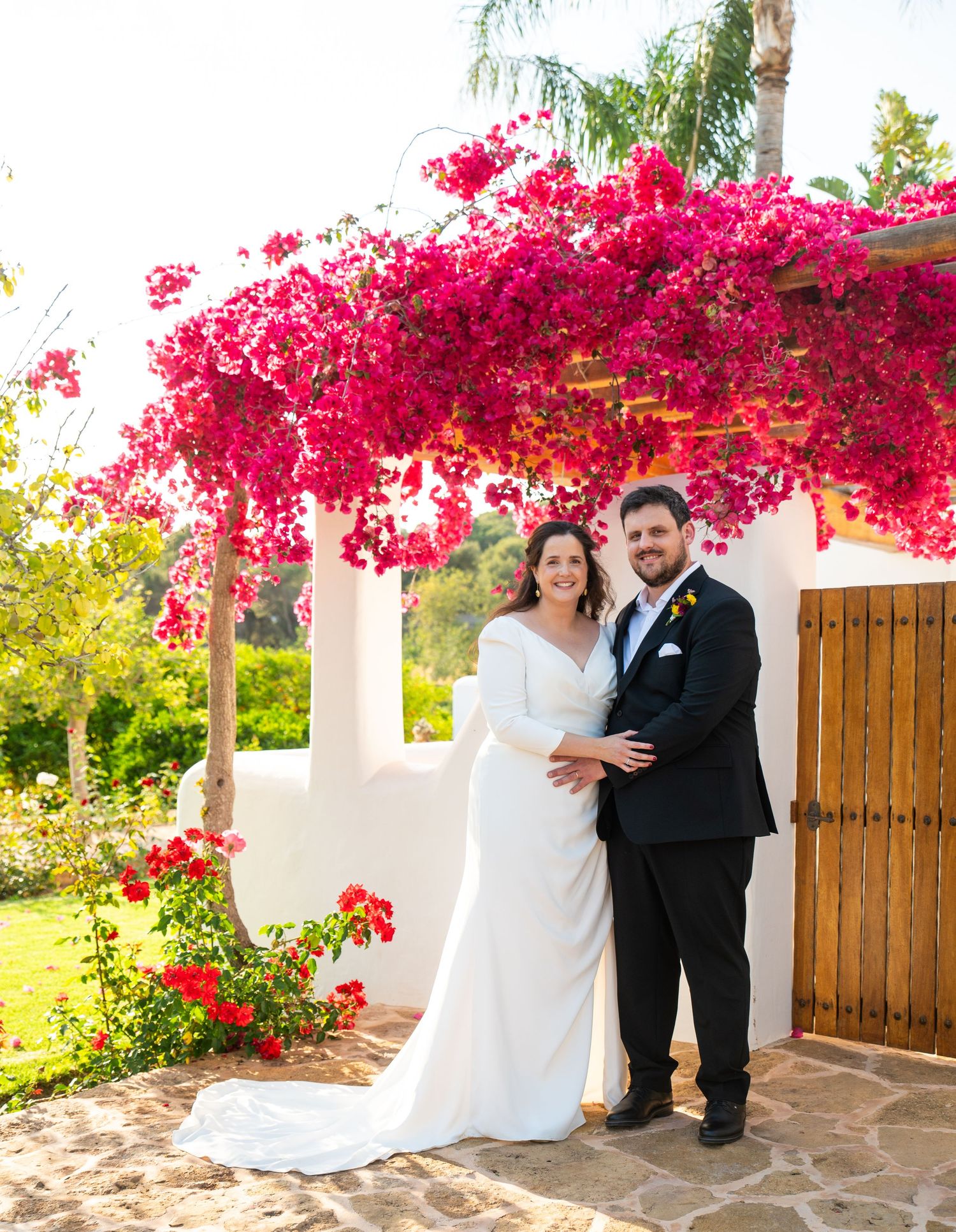Bride and groom pose under vibrant pink bougainvillea at outdoor wedding venue with white stucco walls.