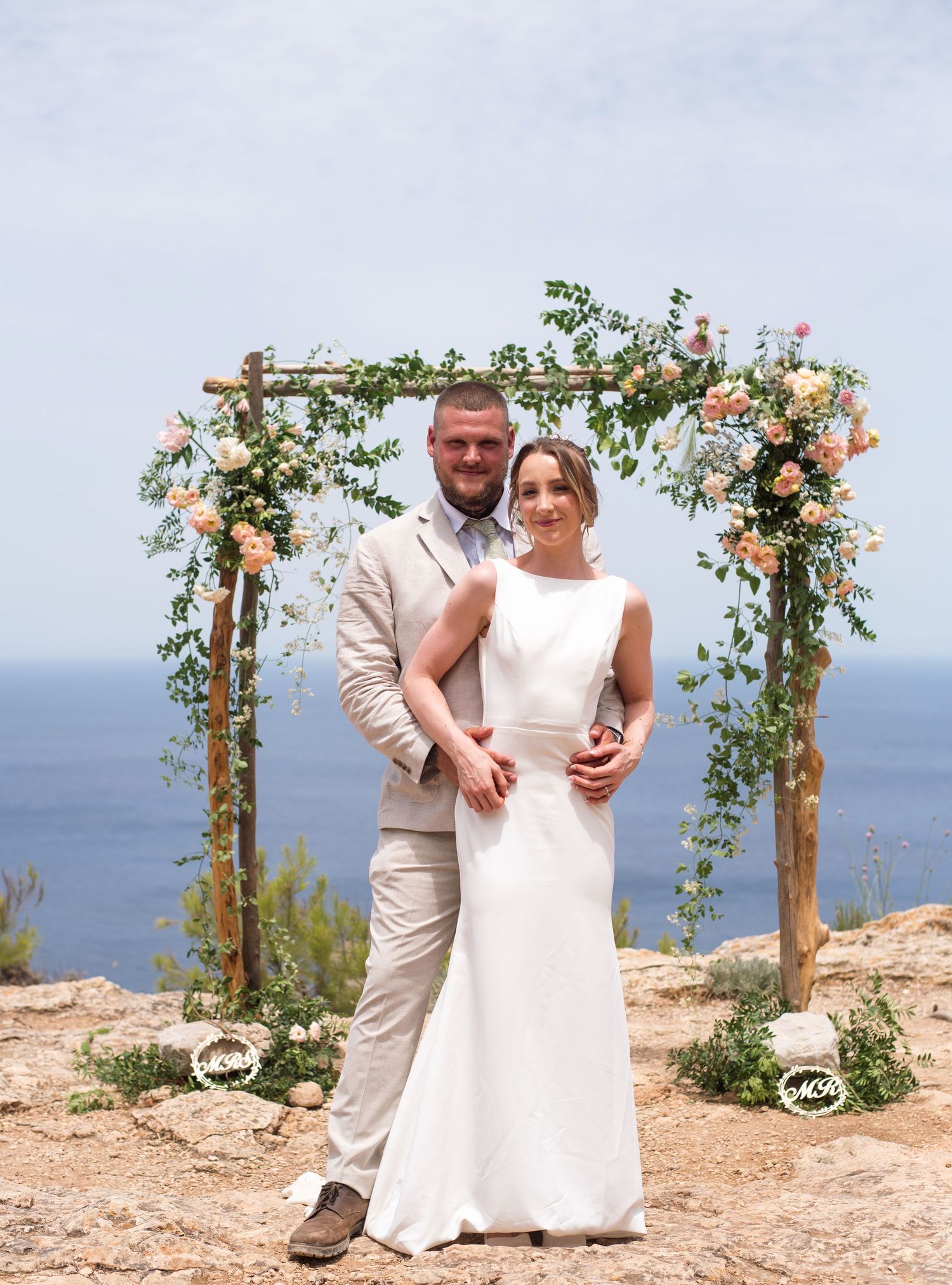 Bride and groom posing under a floral arch with ocean views on their wedding day.