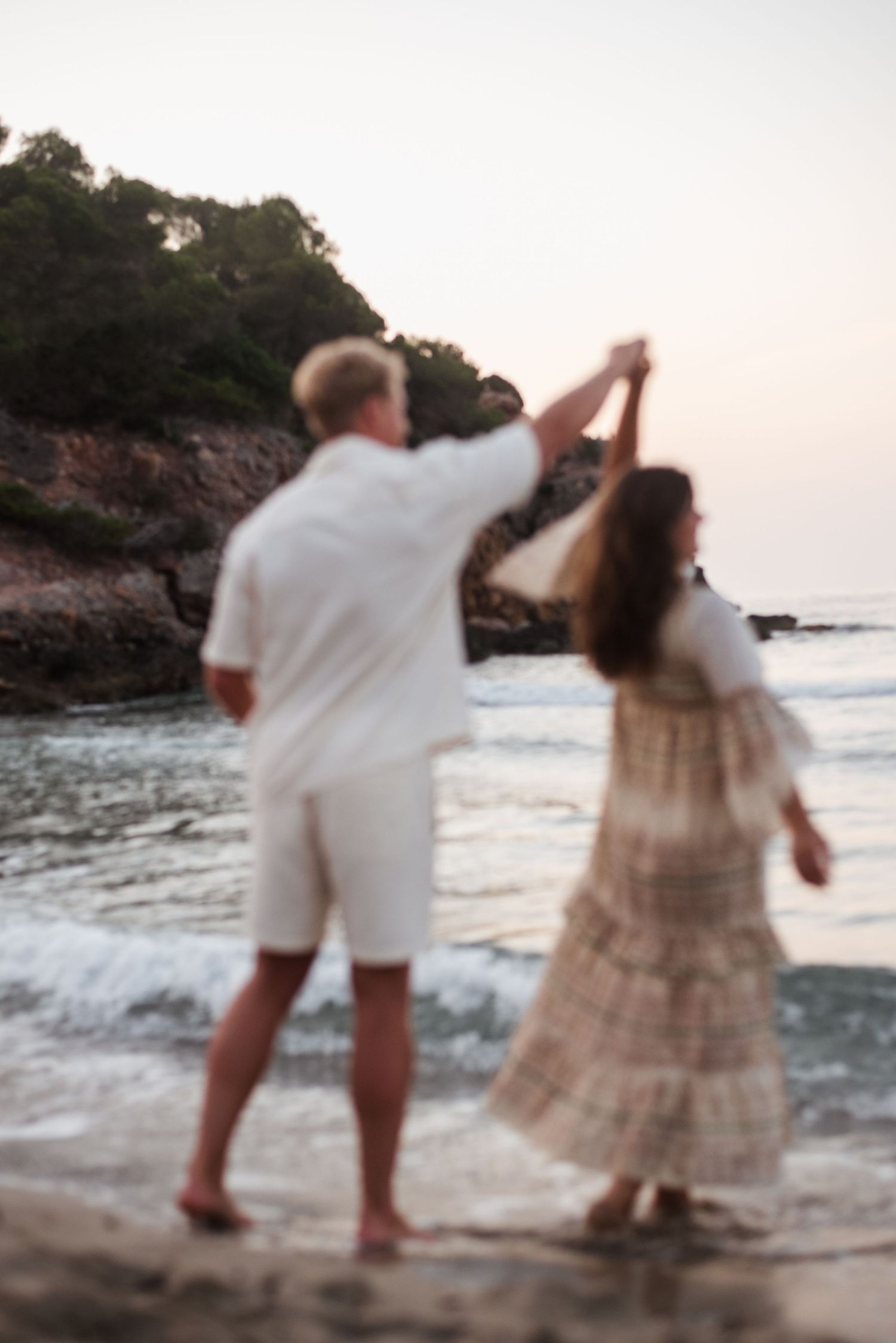 A couple dances barefoot on a beach at sunset, man in white outfit twirling woman in flowing boho dress.