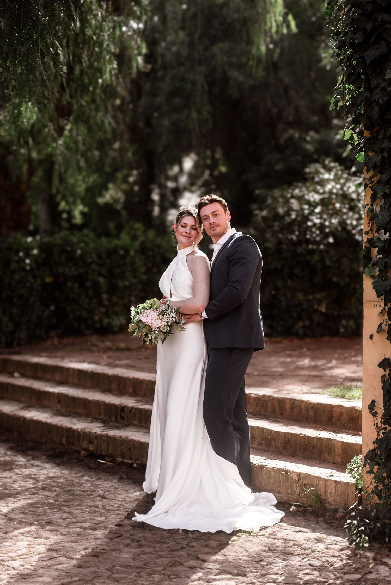Bride and groom pose together in a lush garden setting, she holds a bouquet in a white gown, he wears a dark suit.