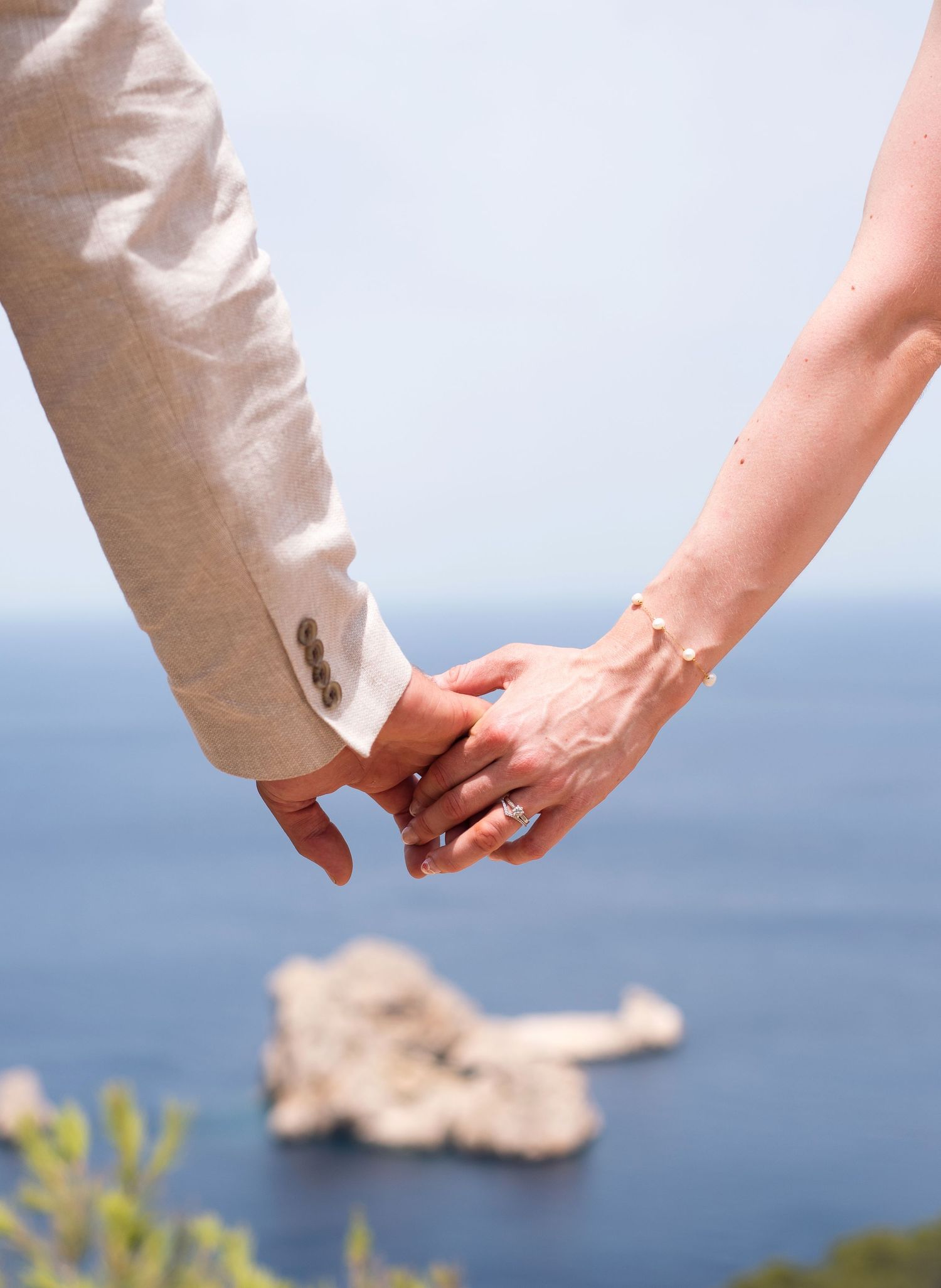 Couple holding hands in wedding attire with a scenic ocean view and rocky island in the background.