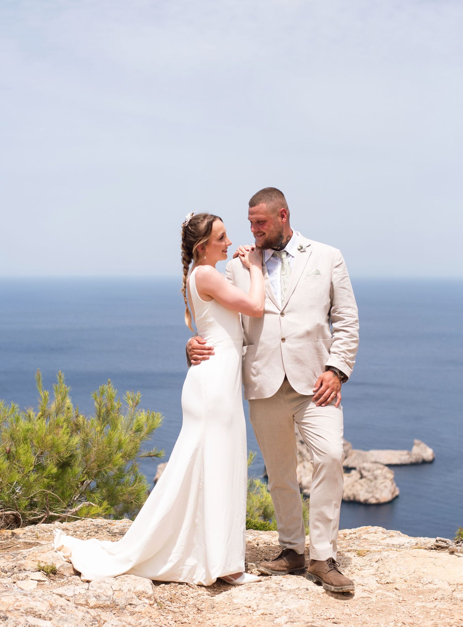 Bride and groom share a romantic moment on a clifftop overlooking the ocean, she in white gown, he in beige suit.