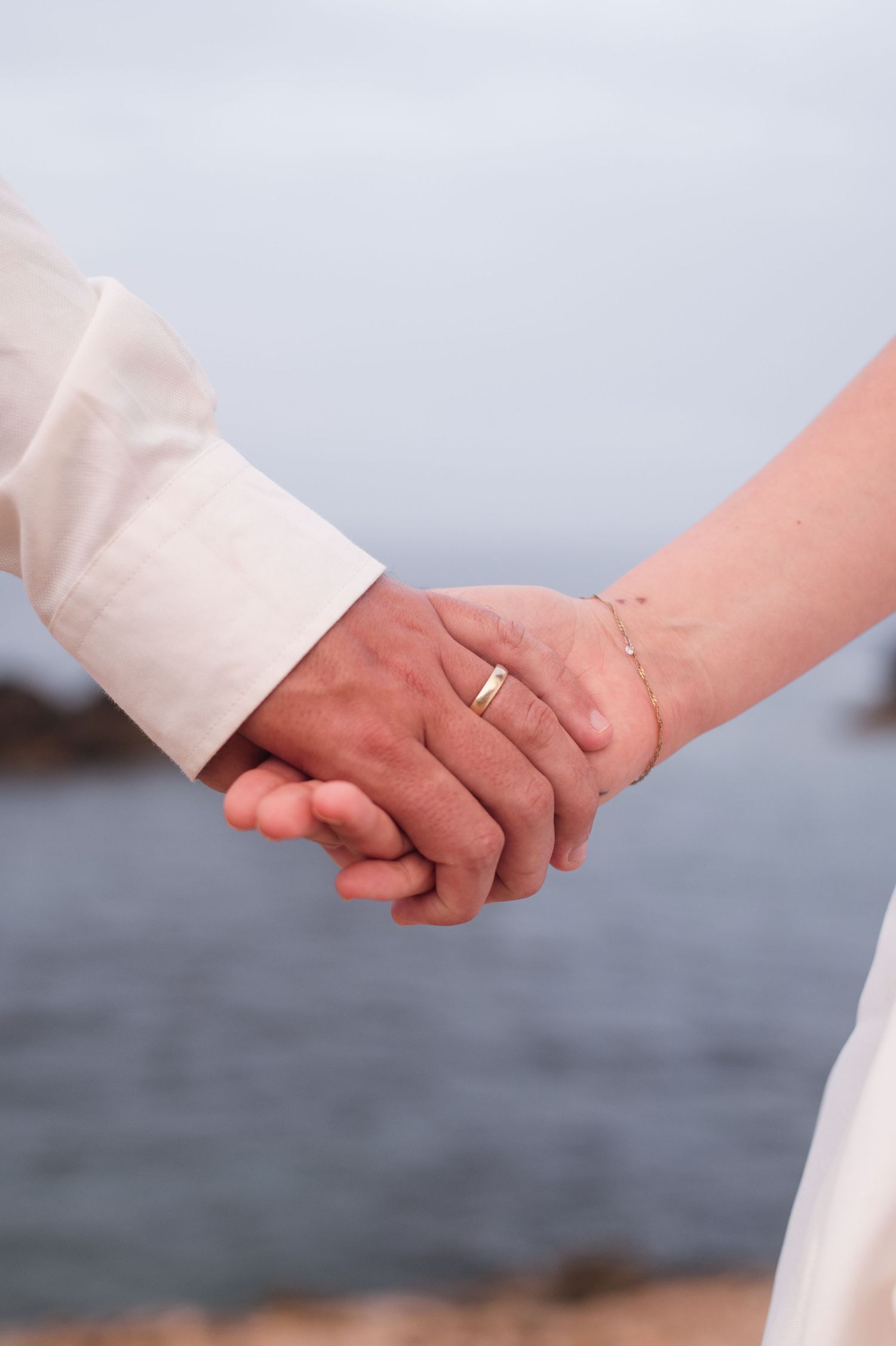 Couple holding hands wearing wedding rings and white attire near ocean backdrop.