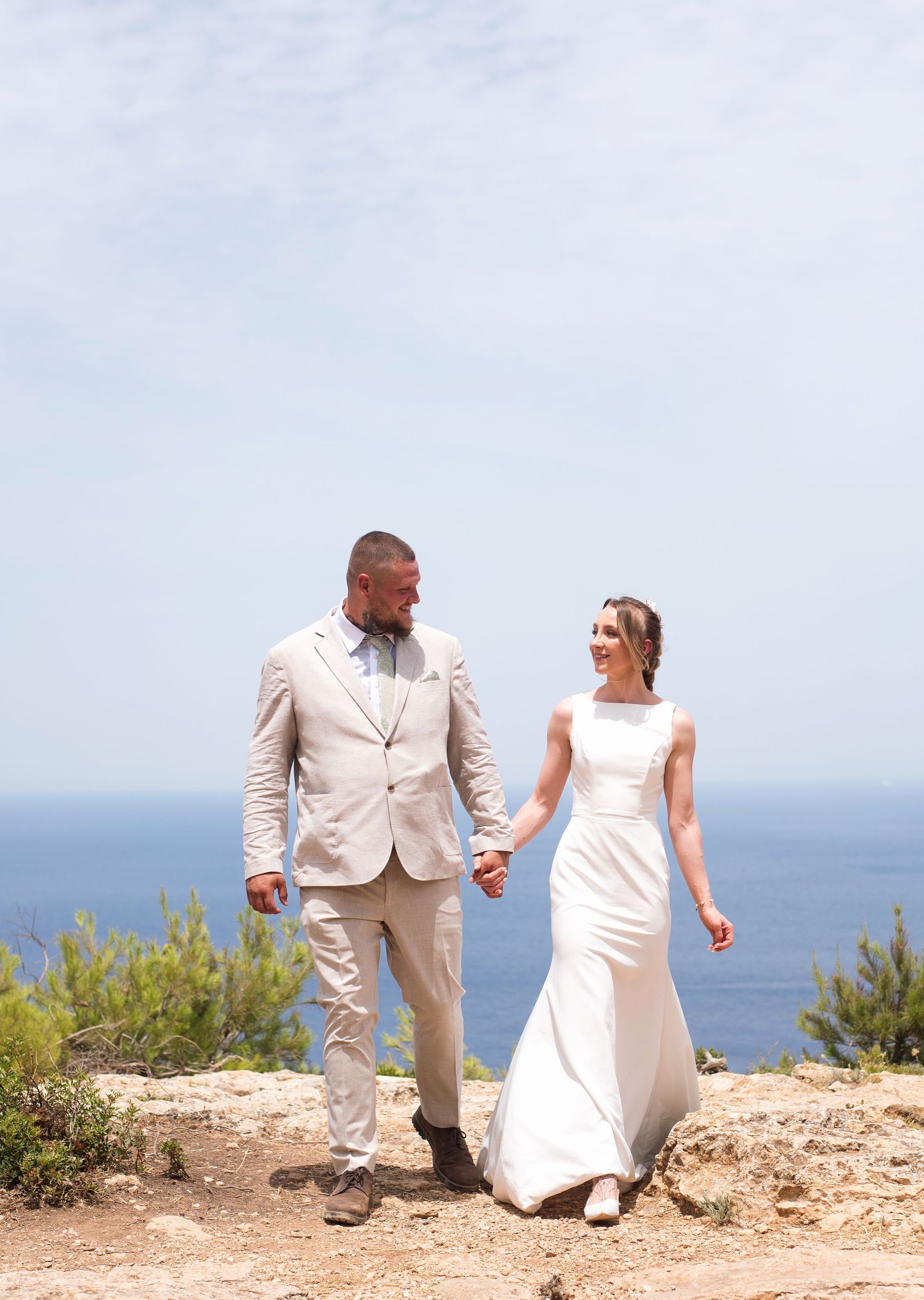 Bride and groom walk hand-in-hand on a coastal cliff, smiling at each other, with ocean views behind them.