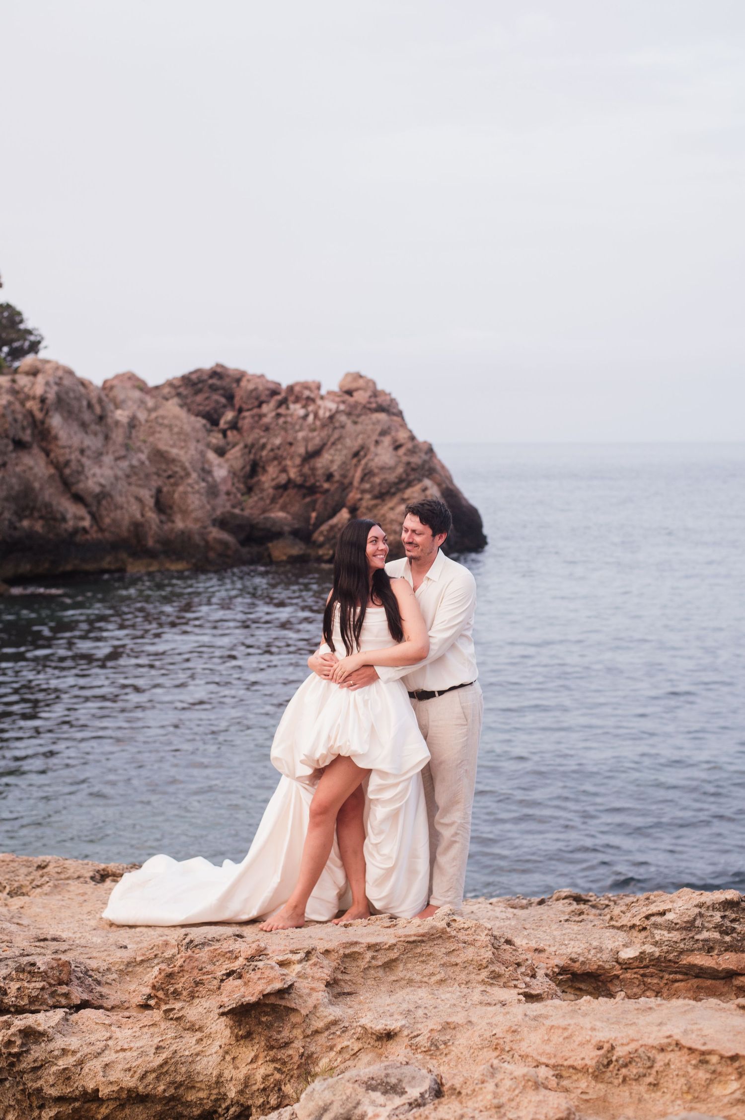 Couple in wedding attire embracing on rocky coastal cliffs with ocean and rock formations in background.