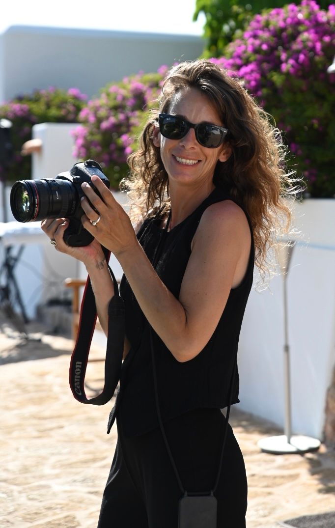 Smiling woman with curly hair and sunglasses holding a Canon camera outdoors near white walls and pink flowers.