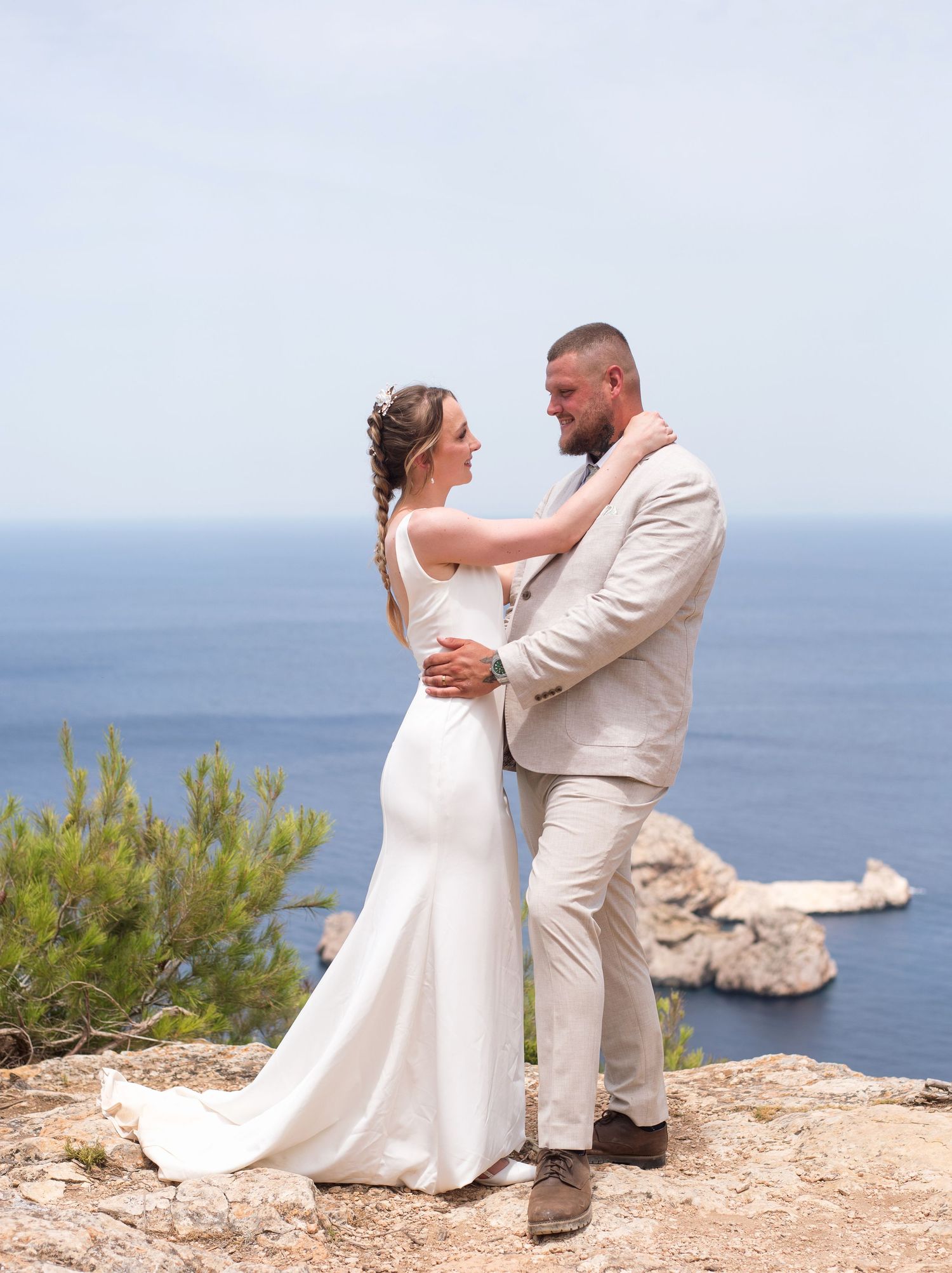 Bride and groom embrace on rocky cliffside overlooking the blue Mediterranean sea on their wedding day.