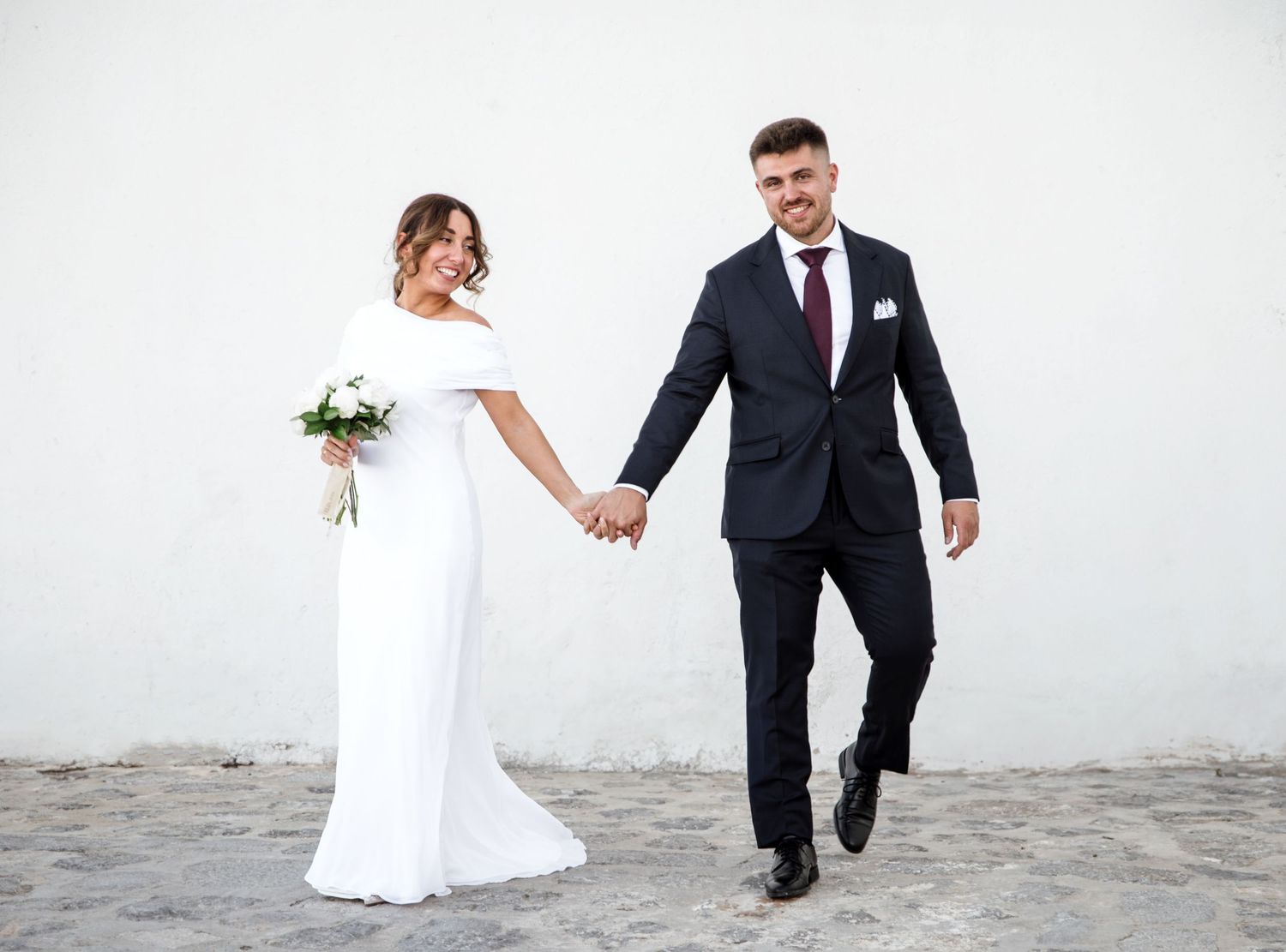 Bride in white gown and groom in navy suit hold hands walking together on their wedding day.