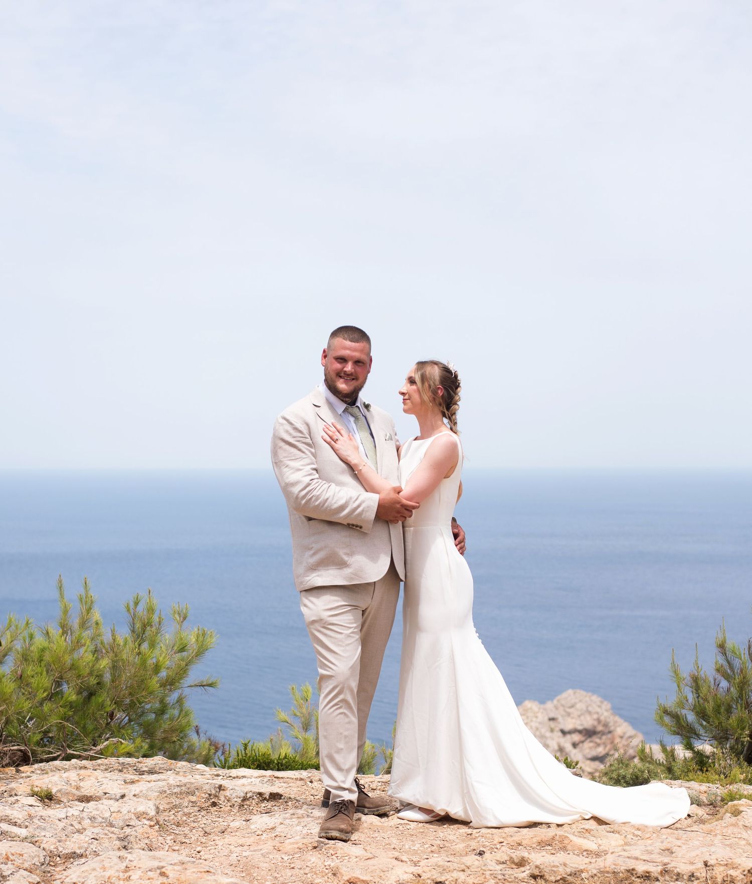 Bride and groom pose together on a rocky coastal cliff with stunning blue ocean views on their wedding day.