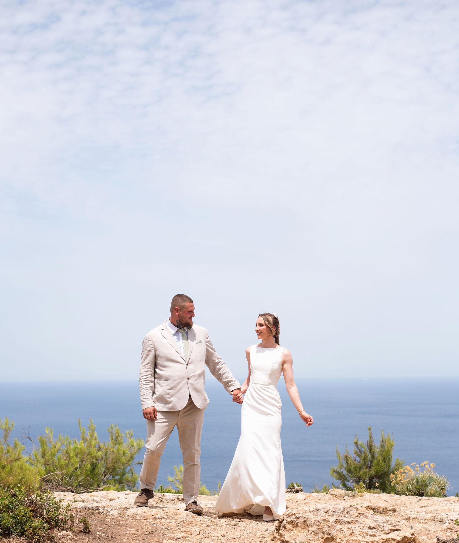 Bride and groom holding hands on a rocky cliff overlooking the blue ocean on their wedding day.