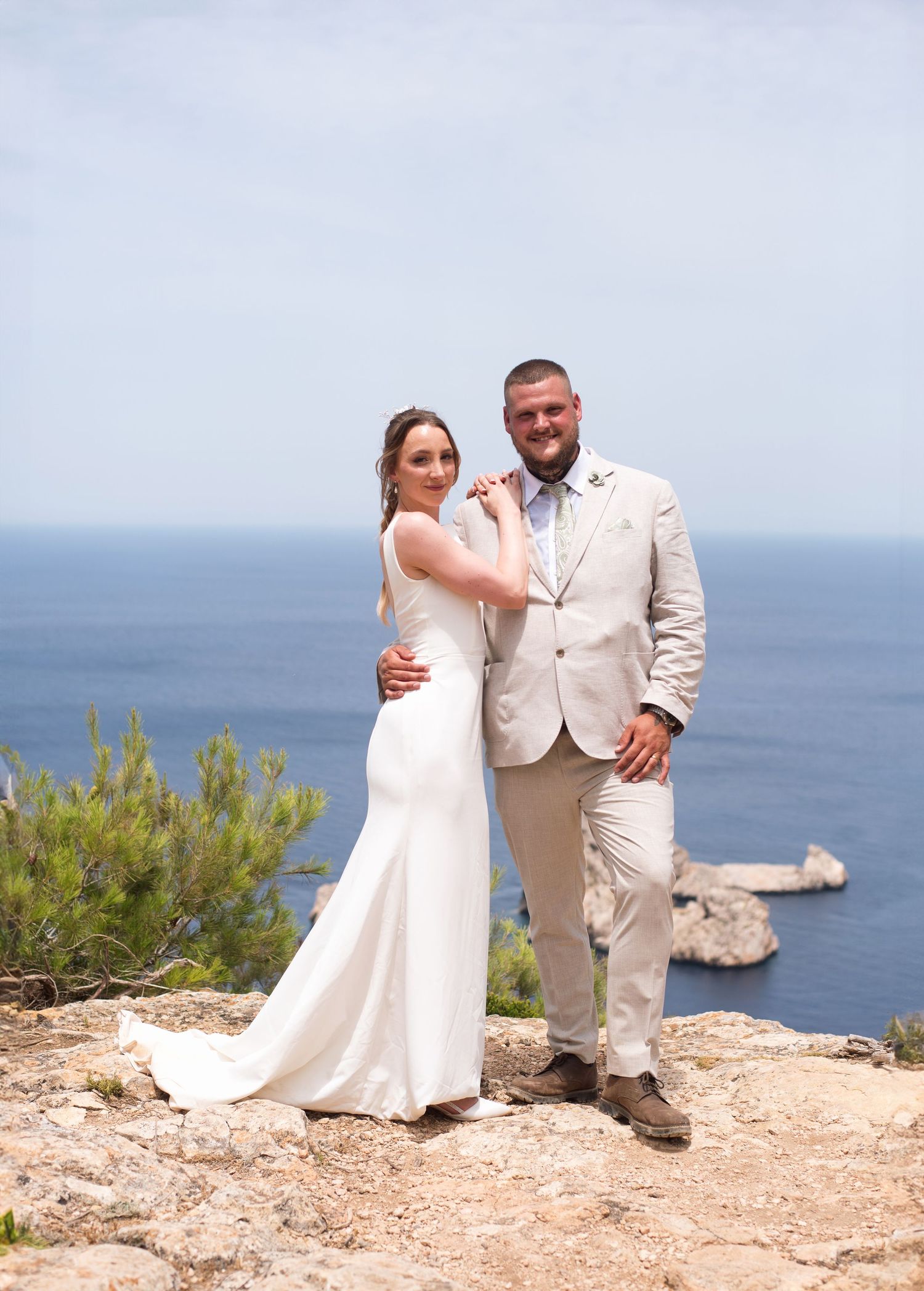 A bride in a white gown and groom in a beige suit pose on a coastal cliff with blue ocean views.